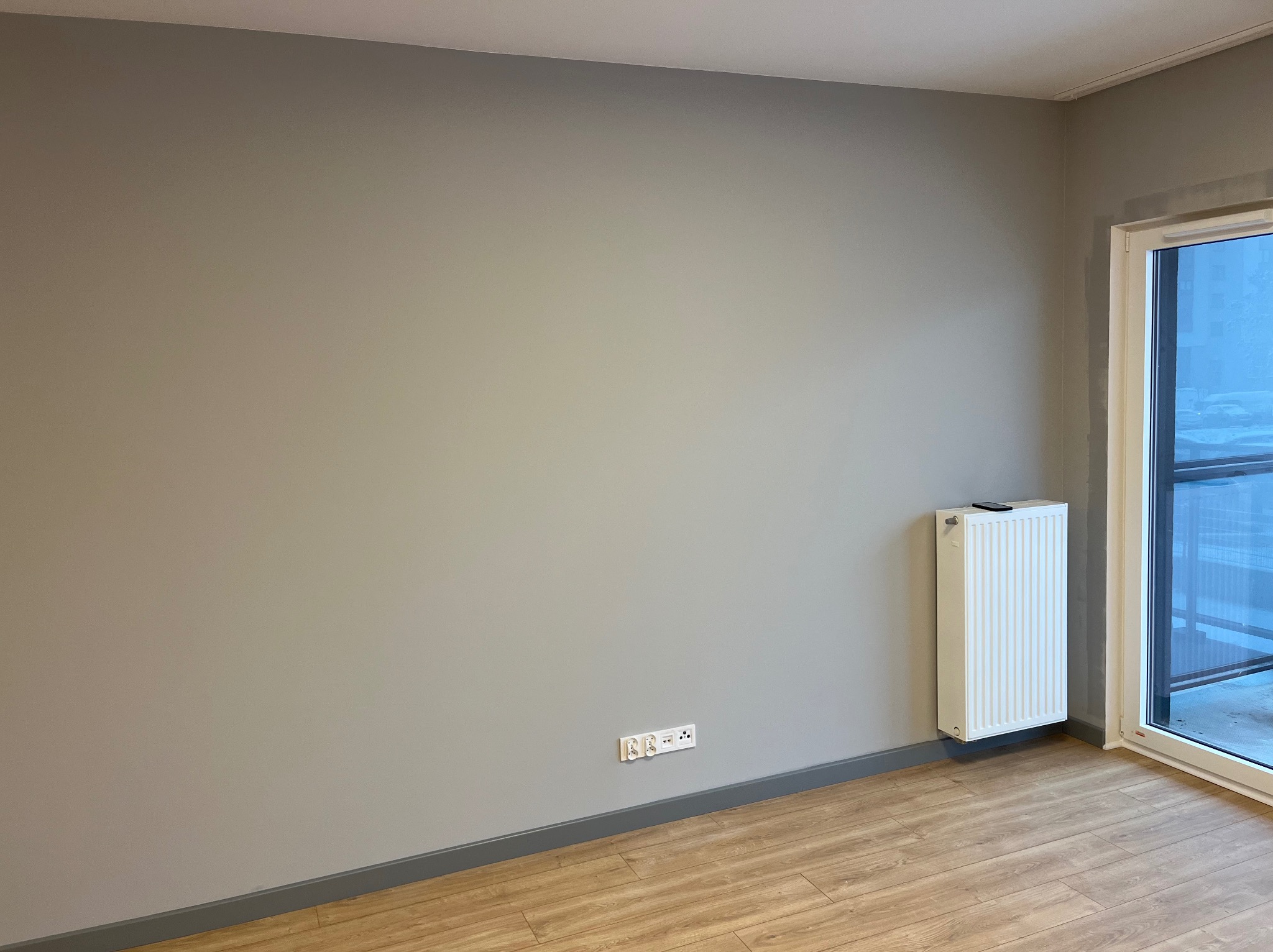 Pomieszczenie po malowaniu ścian na szaro, z drewnopodobną podłogą, białym grzejnikiem i oknem balkonowym. Widoczne gniazdka elektryczne przy podłodze.