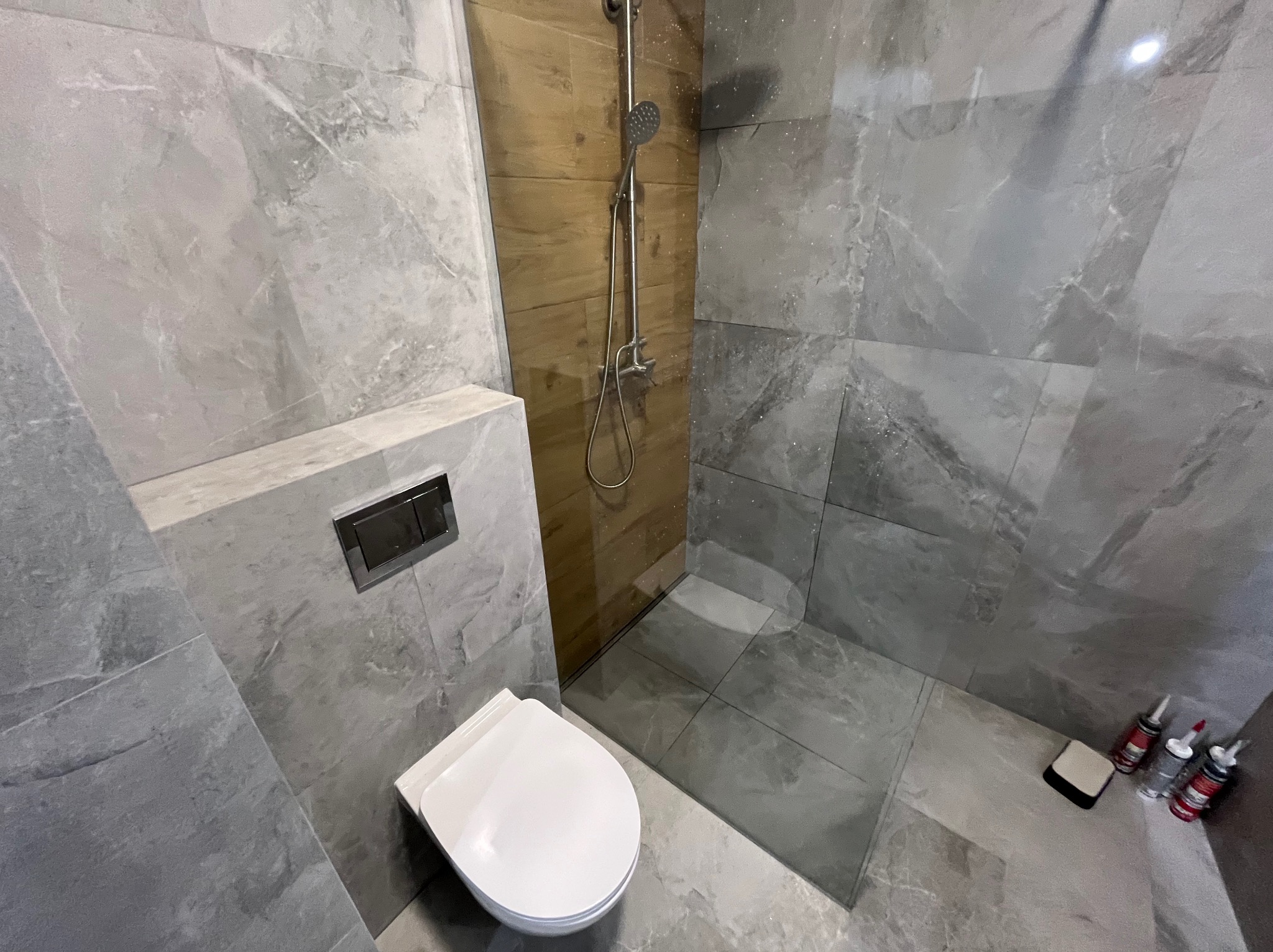 Nowoczesna łazienka z szarymi płytkami imitującymi marmur, kabiną prysznicową typu walk-in i podwieszaną toaletą. Widoczne detale wykończeniowe i armatura.