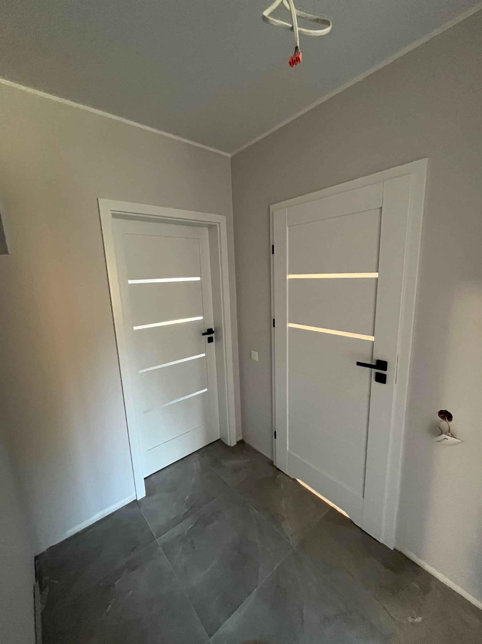 Dwa białe, nowoczesne drzwi z czarnymi klamkami, zamontowane w szarym korytarzu z szarą podłogą. Widoczne gniazdko elektryczne i przewody oświetleniowe.