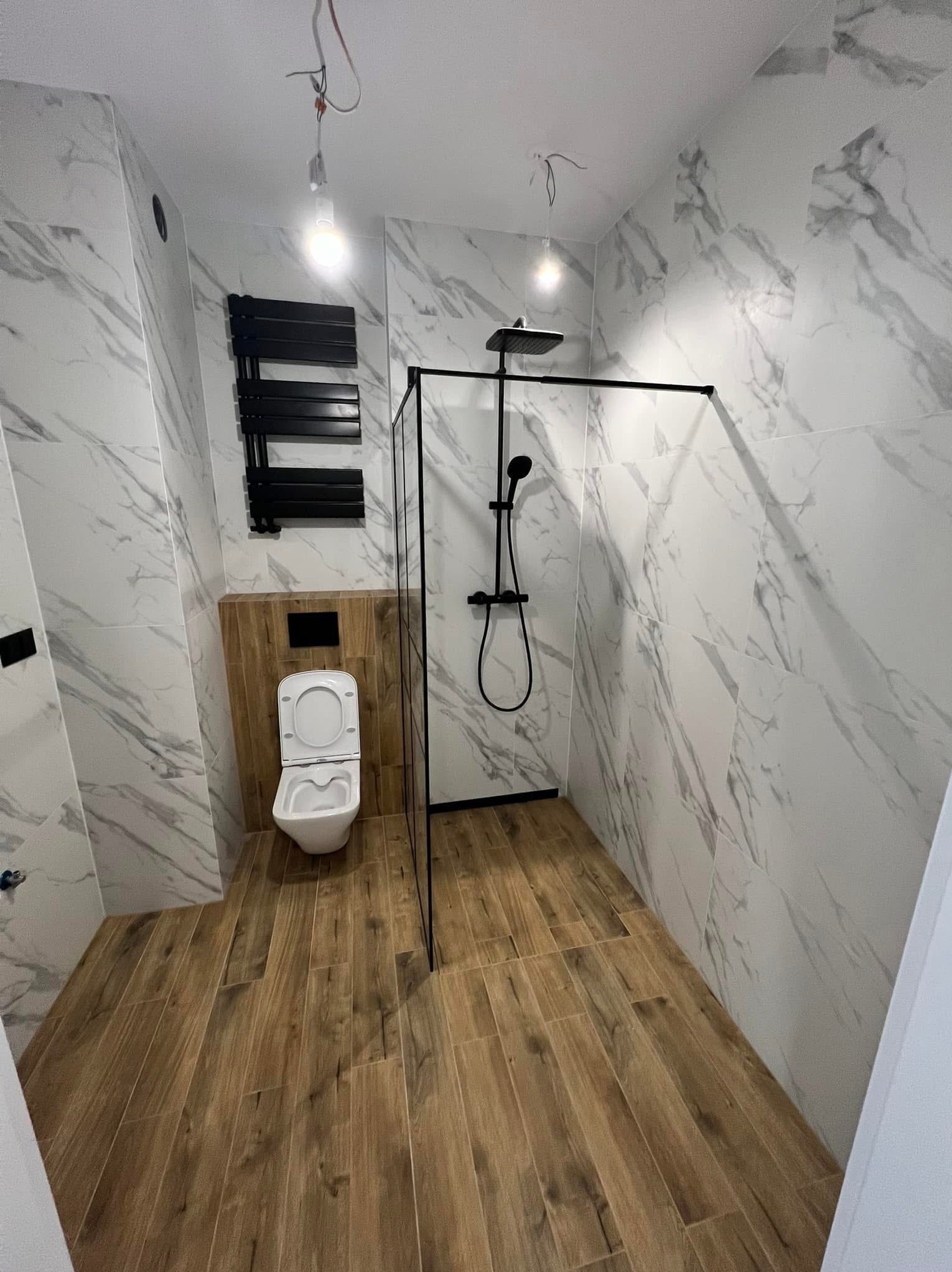 Nowoczesna łazienka z marmurowymi płytkami, drewnopodobną podłogą i czarną armaturą. Widoczny otwarty prysznic i wisząca toaleta. Niewykończone oświetlenie sufitowe.