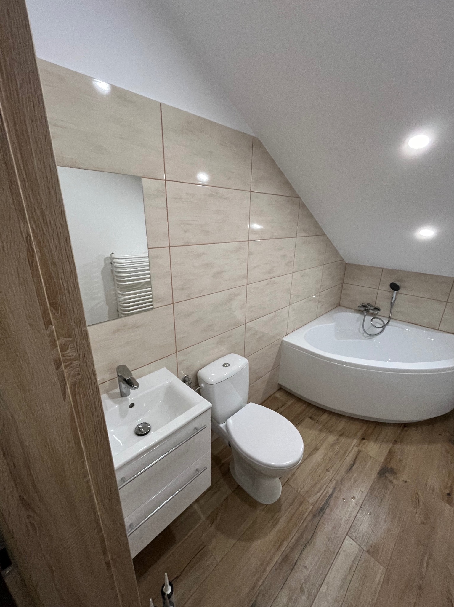 Jasna łazienka na poddaszu z wanną narożną, umywalką z szafką i toaletą. Beżowe płytki na ścianach, drewnopodobne na podłodze. Lustro odbija grzejnik.