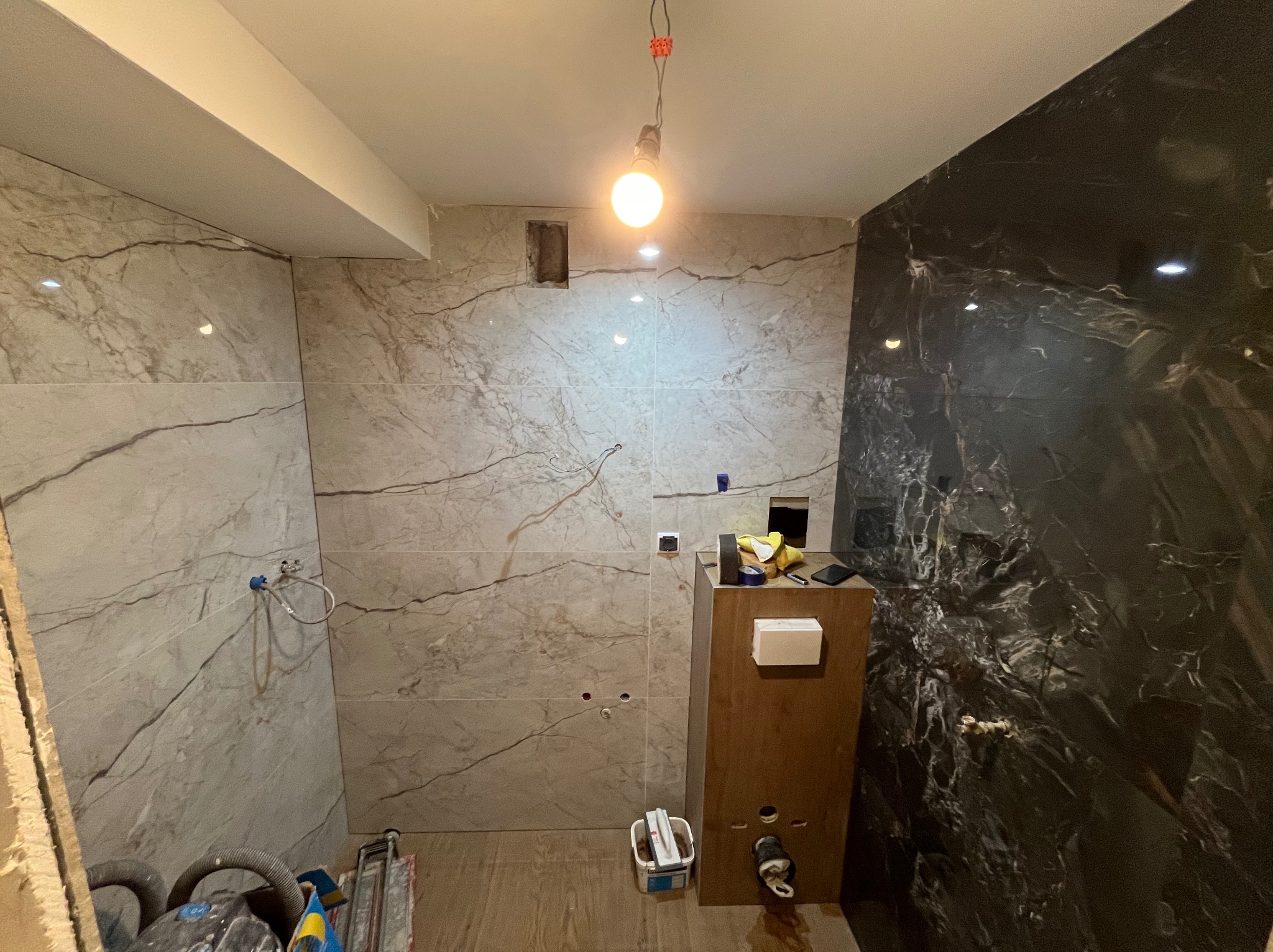 Łazienka w trakcie wykończenia: marmurowe płytki w odcieniach szarości i beżu na ścianach, zabudowa WC, widoczne narzędzia i akcesoria glazurnicze, surowa żarówka.