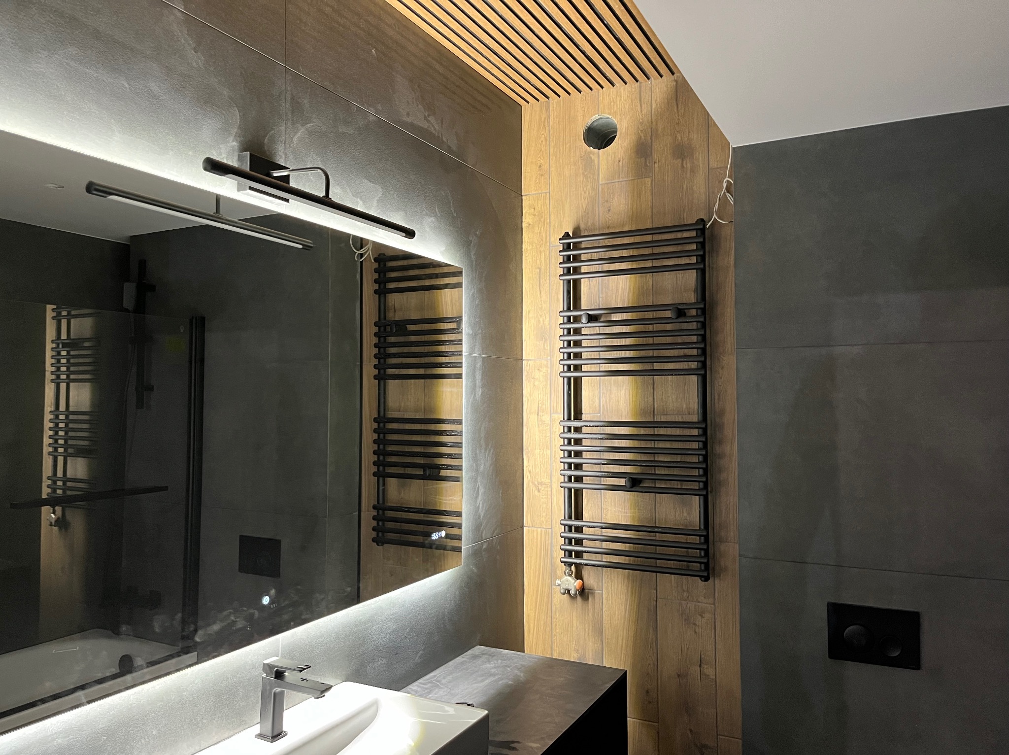 Nowoczesna łazienka w Dąbrowie Górniczej: szare płytki, drewnopodobna okładzina, czarny grzejnik drabinkowy i podświetlane lustro z umywalką.