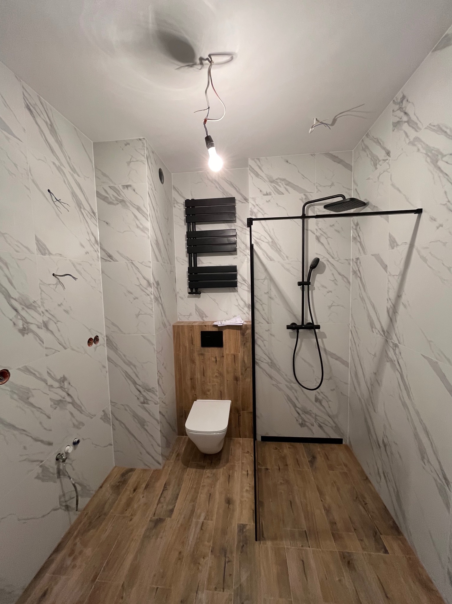 Nowoczesna łazienka w trakcie wykańczania: białe marmurowe płytki, drewnopodobna podłoga, czarna kabina prysznicowa i grzejnik. Widoczne niedokończone instalacje.