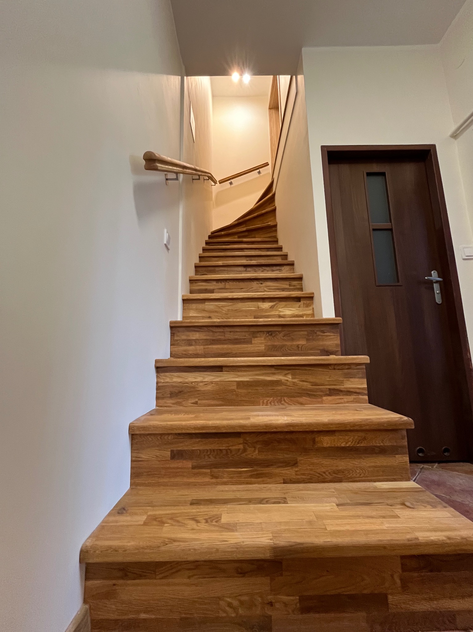 Drewniane schody z poręczą, prowadzące do góry, obok ciemnych drzwi. Widok z dołu, podkreślający solidne wykonanie i naturalny odcień drewna.