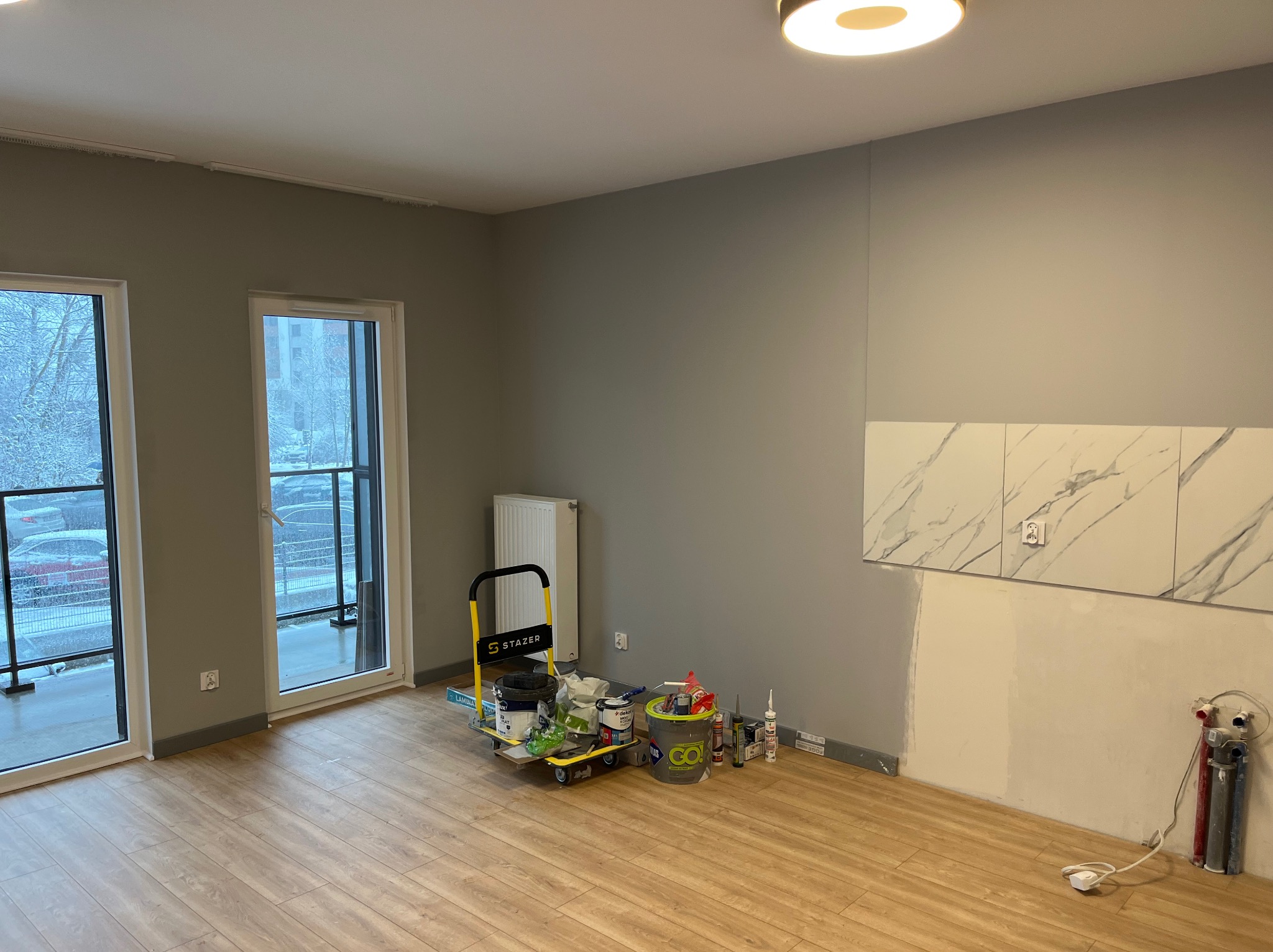 Wnętrze w trakcie malowania: szare ściany, panele podłogowe, wózek z farbami i akcesoriami malarskimi, częściowo wykończona ściana z marmurowymi płytkami. Widoczne okna balkonowe.