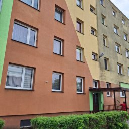 Clean-Mar Wiesław Śliwa - Wielopiętrowy budynek po częściowym myciu elewacji, z widoczną różnicą w kolorze między umytą a brudną powierzchnią. Osoba w czerwonej kurtce myje elewację lancą.