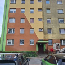 Clean-Mar Wiesław Śliwa - Wielobarwny blok po częściowym myciu elewacji. Widoczny kontrast czystej, żółtej ściany z brudną, szarą. Pracownik na podnośniku kontynuuje prace.