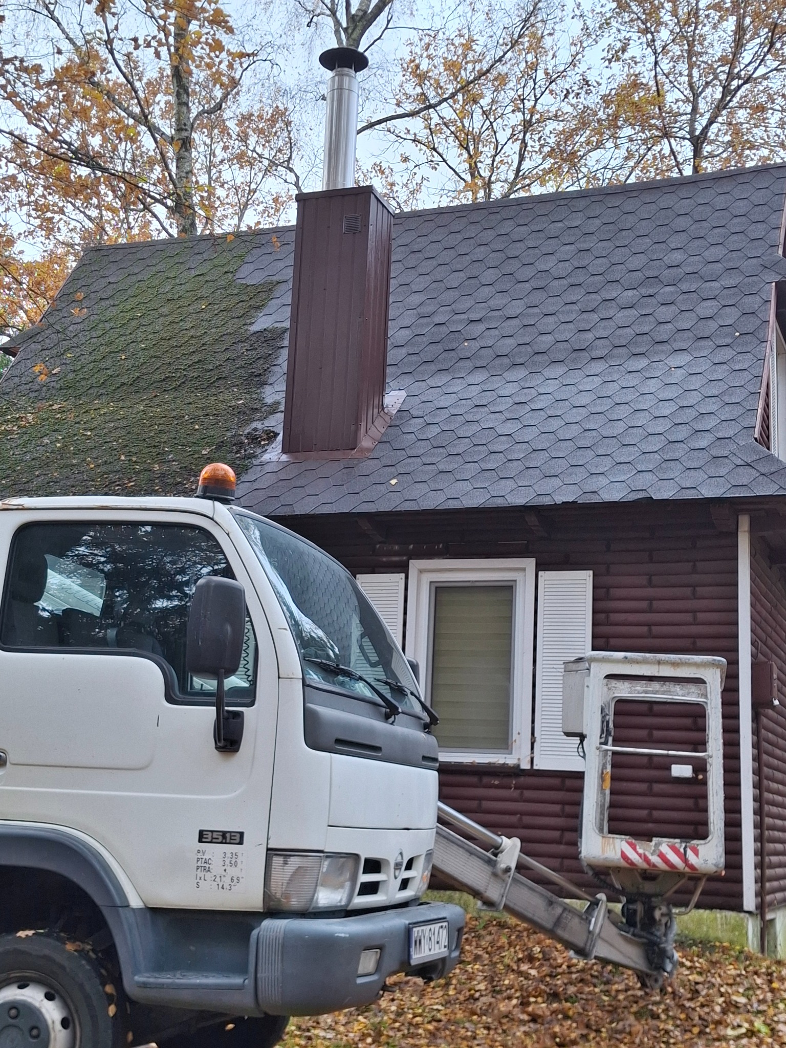 Ciężarówka z podnośnikiem koszowym na tle domu z dachem pokrytym częściowo mchem. Widoczny komin i jesienne drzewa. Prace na wysokości.