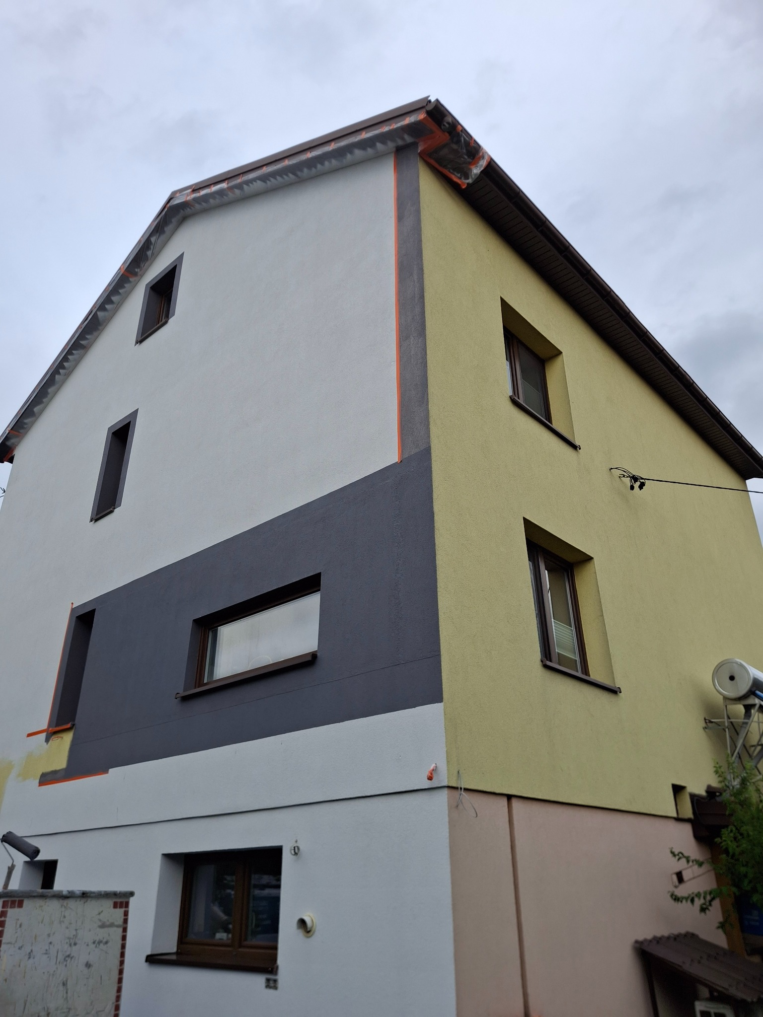 Wielobarwna elewacja domu w trakcie malowania. Widoczne fragmenty w kolorze białym, szarym i żółtym. Przy ścianie stoi wałek malarski. Ujęcie z dołu, pochmurne niebo.