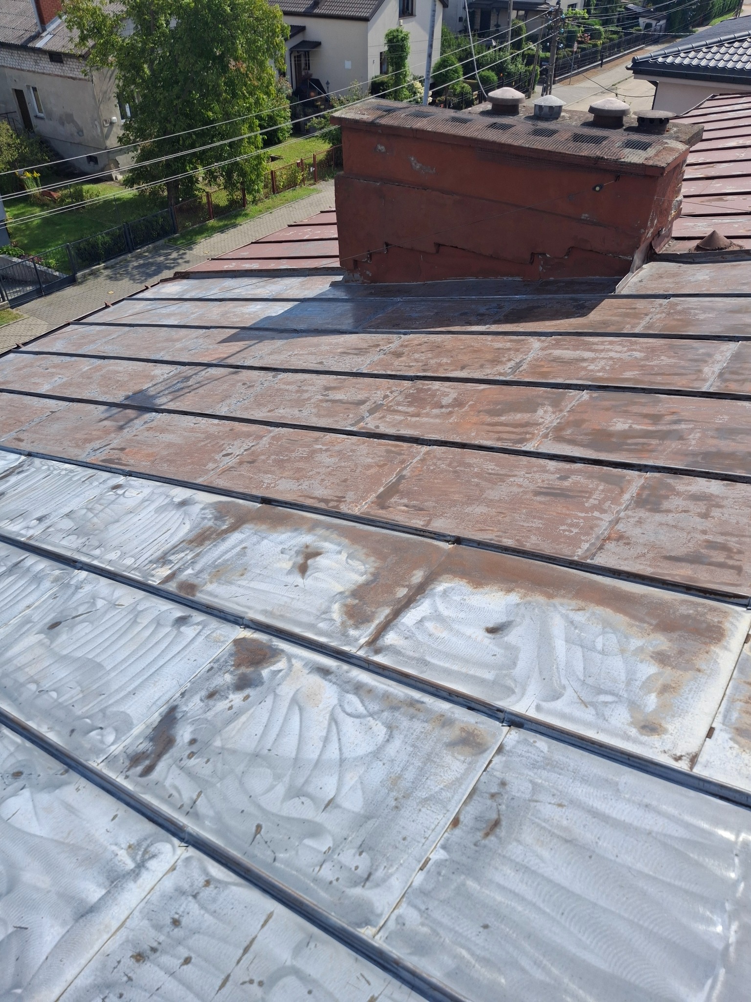 Częściowo wyczyszczony dach z blachy, widoczny komin i fragmenty zabudowy w tle. Kontrast między czystą a zabrudzoną powierzchnią blachy.