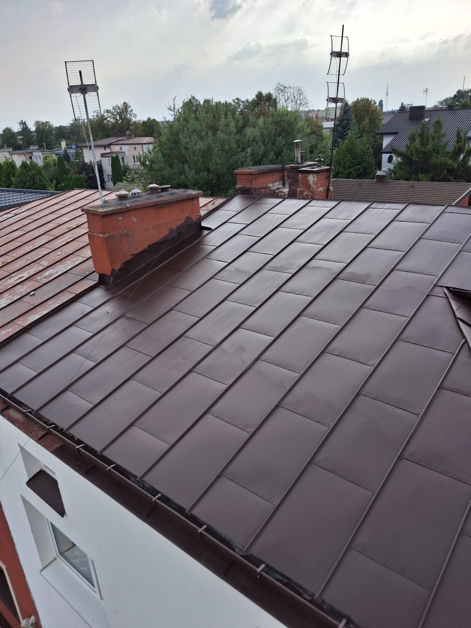 Ciemnobrązowy dach z blachy na budynku z kominem i antenami, widok z góry. Część dachu jest stara, z widocznymi śladami użytkowania. Tło stanowią drzewa i zabudowania.