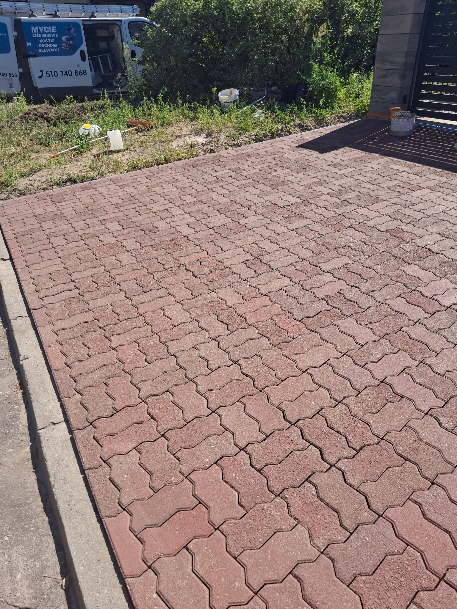 Czerwona kostka brukowa ułożona na podjeździe, widoczny fragment samochodu z napisem 'Mycie ciśnieniowe', narzędzia i wiadro. Słoneczny dzień.