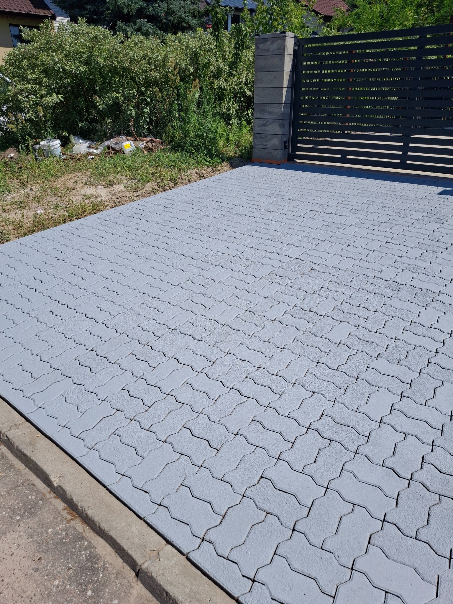 Świeżo pomalowana na szaro kostka brukowa przed posesją z czarną bramą, widoczne ślady farby na fugach. W tle zieleń i zabudowania.