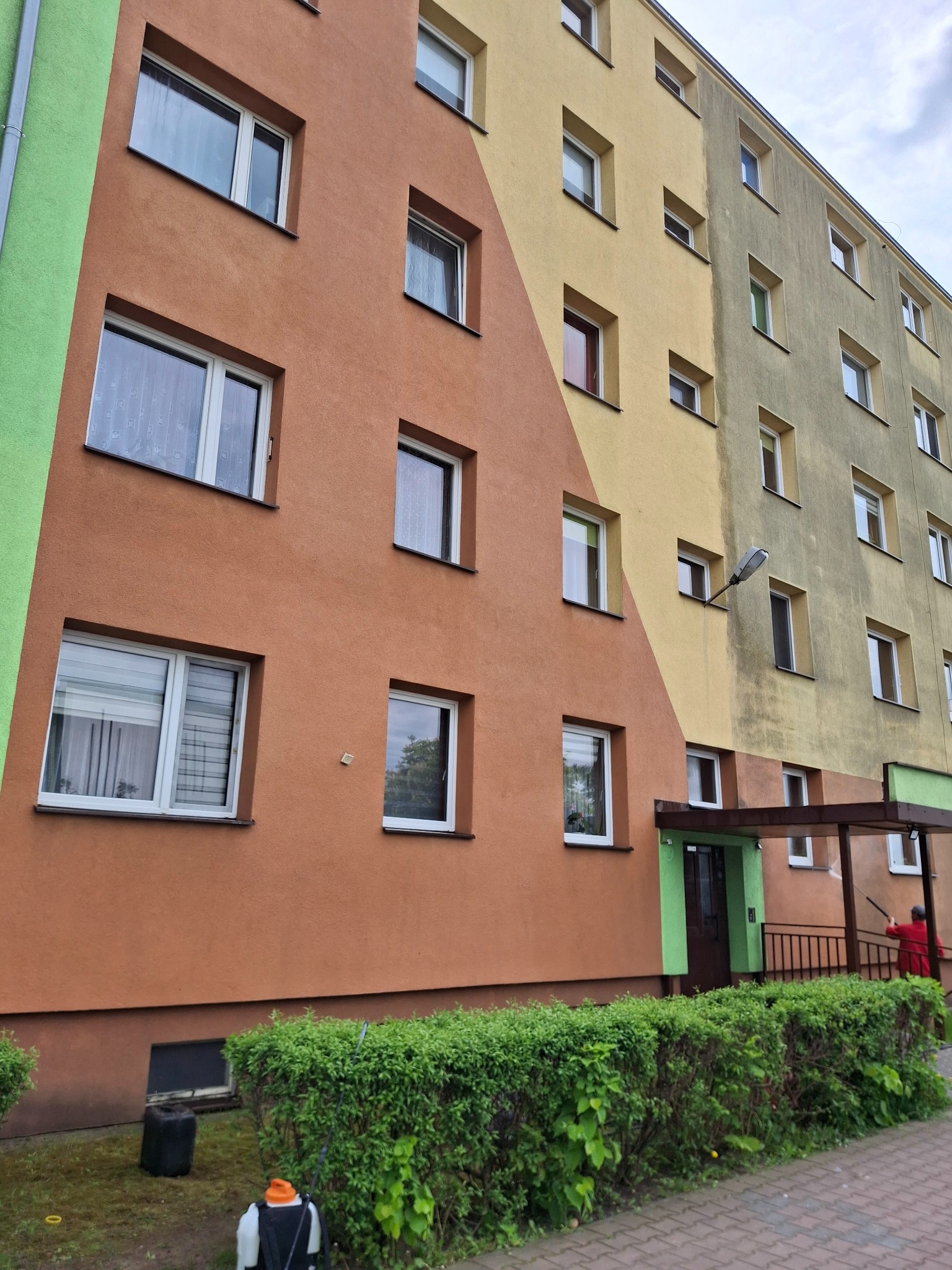 Wielopiętrowy budynek po częściowym myciu elewacji, z widoczną różnicą w kolorze między umytą a brudną powierzchnią. Osoba w czerwonej kurtce myje elewację lancą.