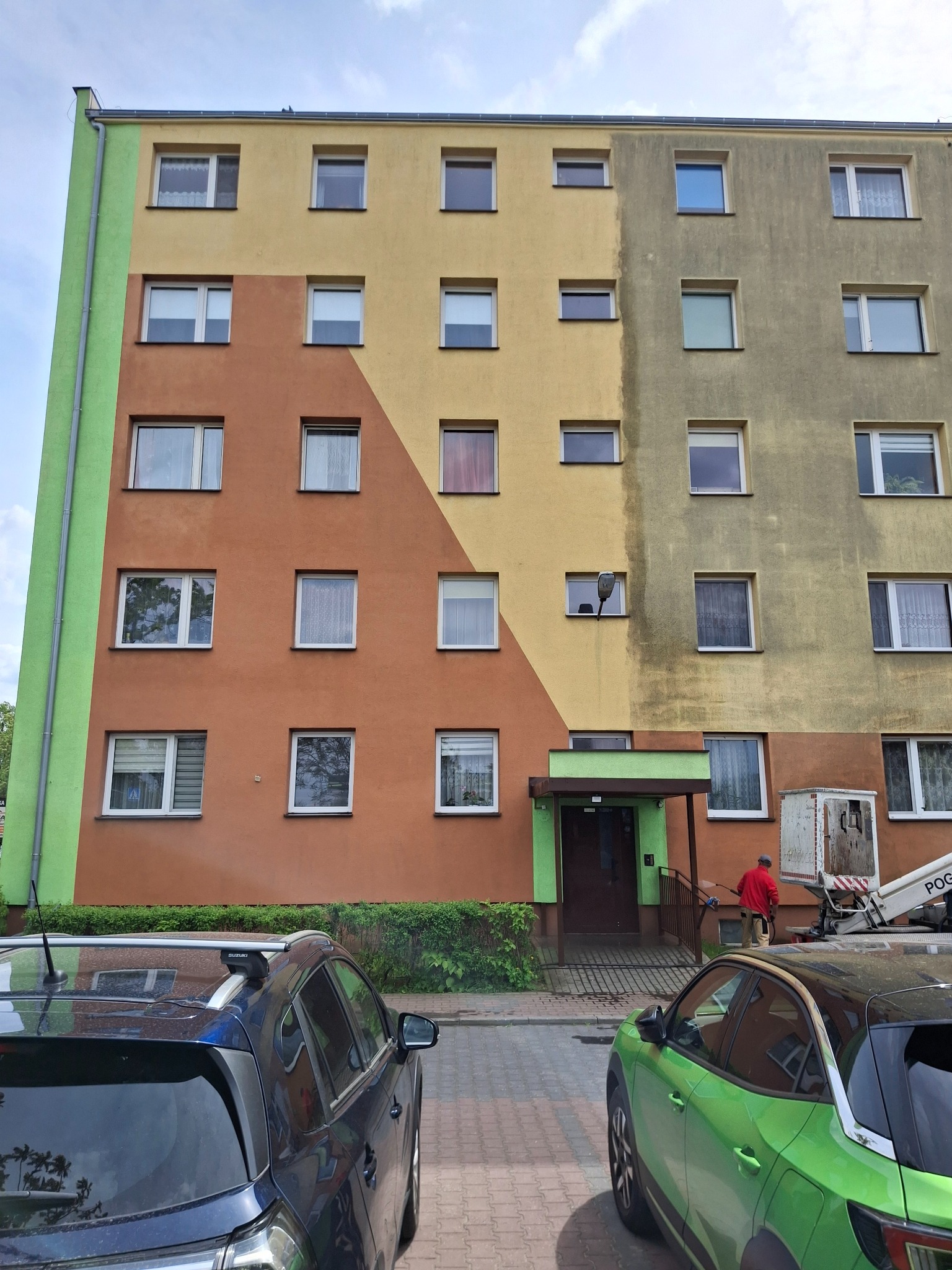 Wielobarwny blok po częściowym myciu elewacji. Widoczny kontrast czystej, żółtej ściany z brudną, szarą. Pracownik na podnośniku kontynuuje prace.