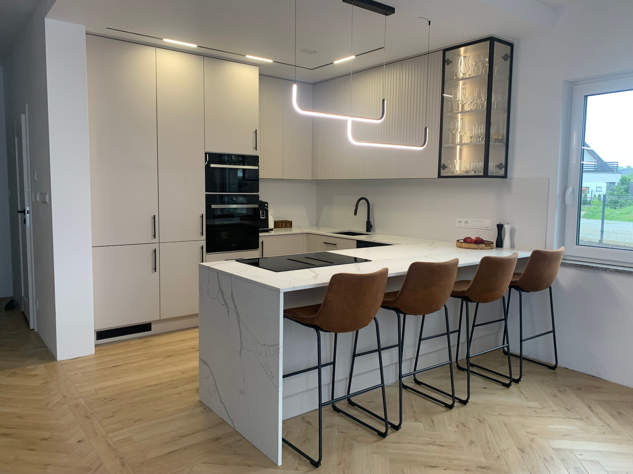 Nowoczesna kuchnia z wyspą i hokerami. Jasne fronty szafek, marmurowy blat wyspy, parkiet w jodełkę. Oświetlenie LED i przeszklona witryna z zastawą.