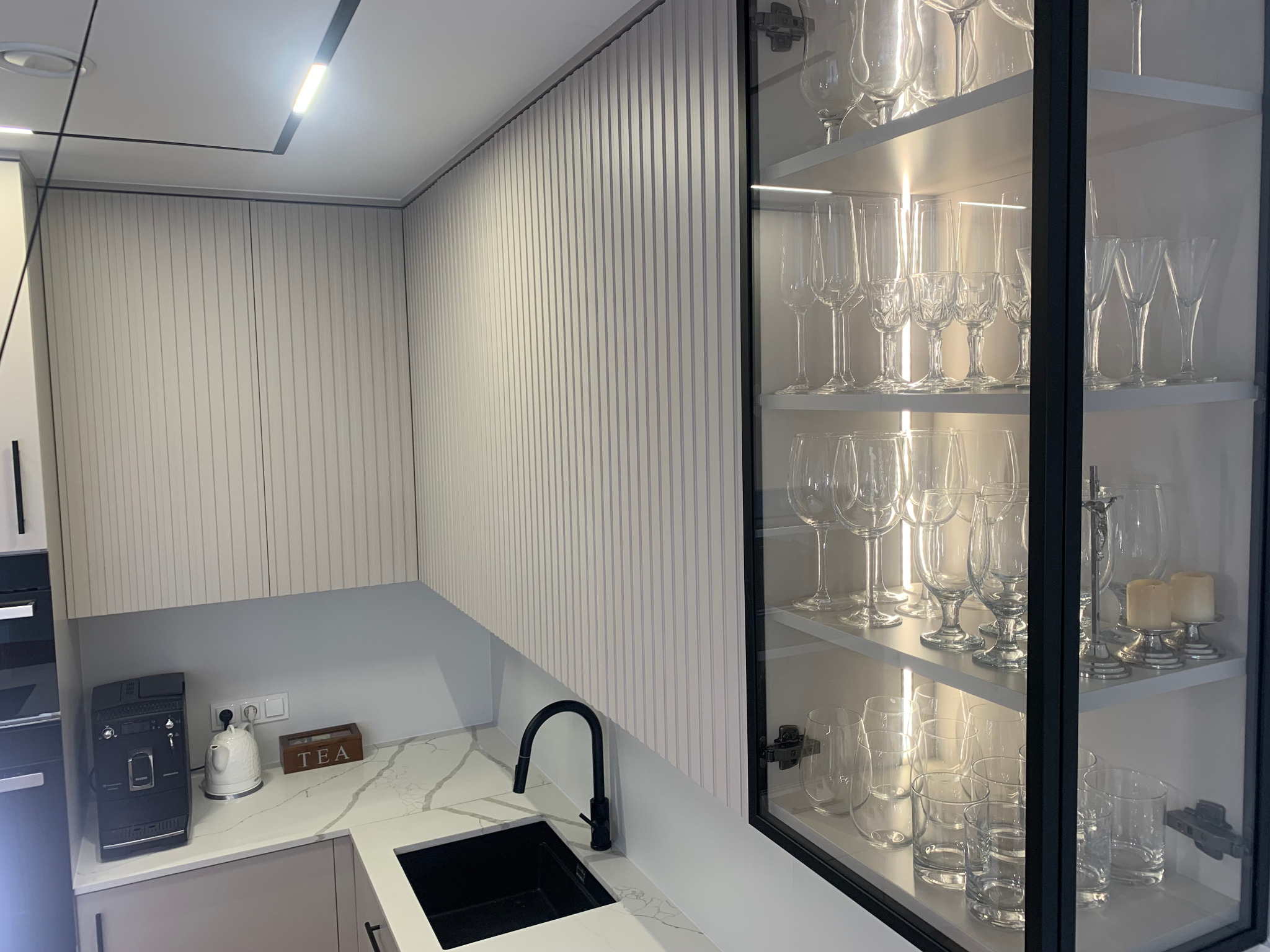Nowoczesna kuchnia: białe, ryflowane szafki wiszące, czarny zlew i przeszklona witryna z kieliszkami, oświetlona LED. Minimalistyczny design.
