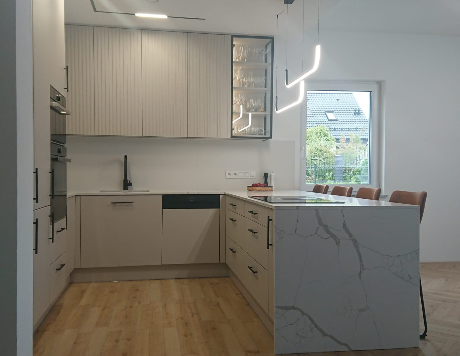 Jasna kuchnia z wyspą i oświetleniem LED. Fronty szafek z subtelnym frezem, czarne uchwyty. Na wyspie płyta grzewcza. W tle okno z widokiem na zieleń.