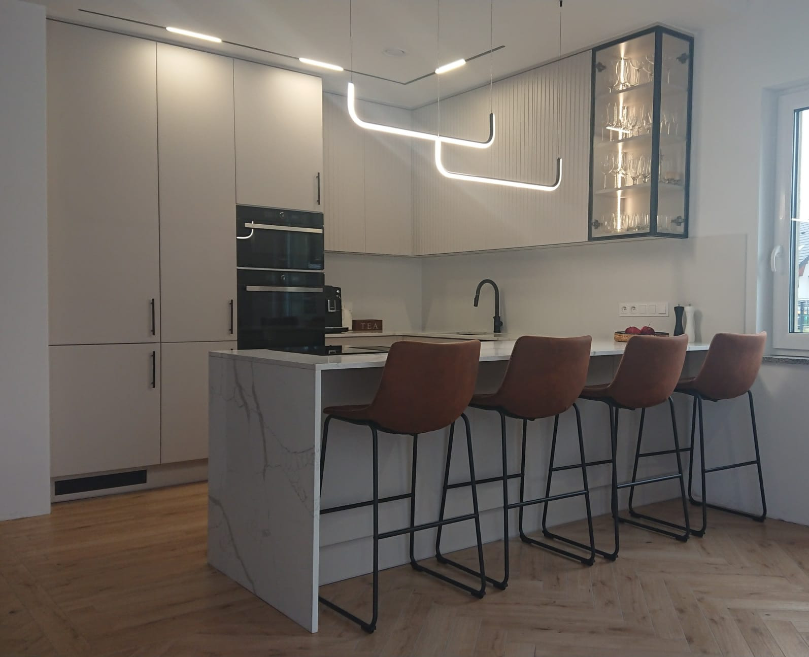 Nowoczesna kuchnia z wyspą i czterema krzesłami barowymi. Białe szafki, marmurowy blat wyspy, parkiet w jodełkę i designerskie oświetlenie nadają wnętrzu elegancji.