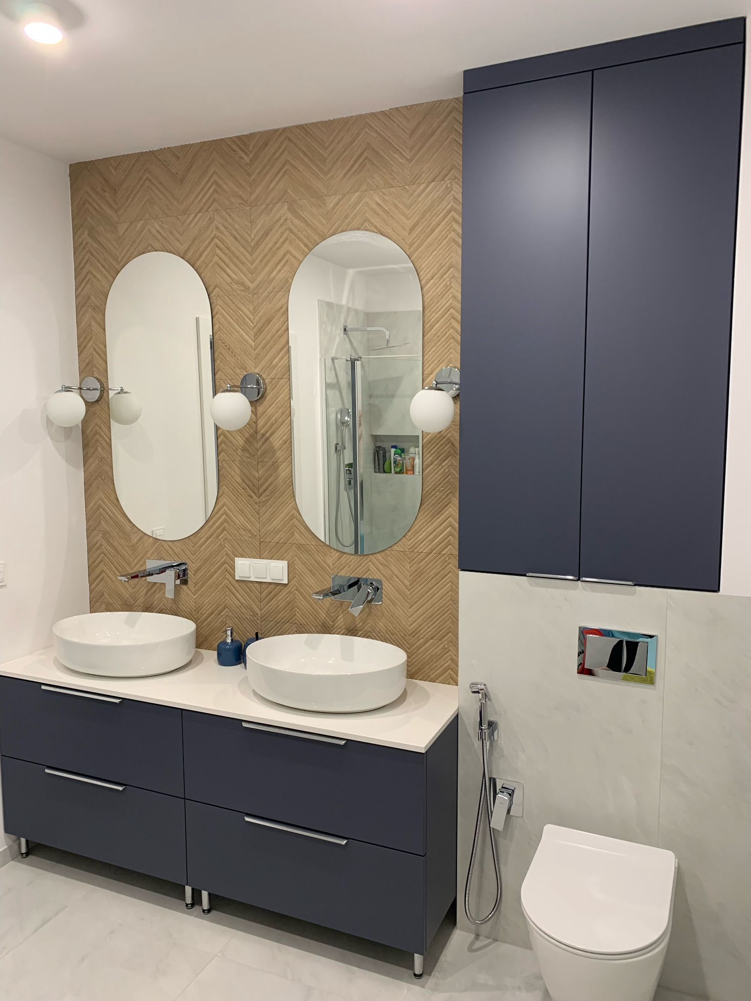 Nowoczesna łazienka z dwoma umywalkami nablatowymi, szafką w kolorze granatowym i lustrami o owalnym kształcie. Na ścianie płytki imitujące drewno ułożone w jodełkę.