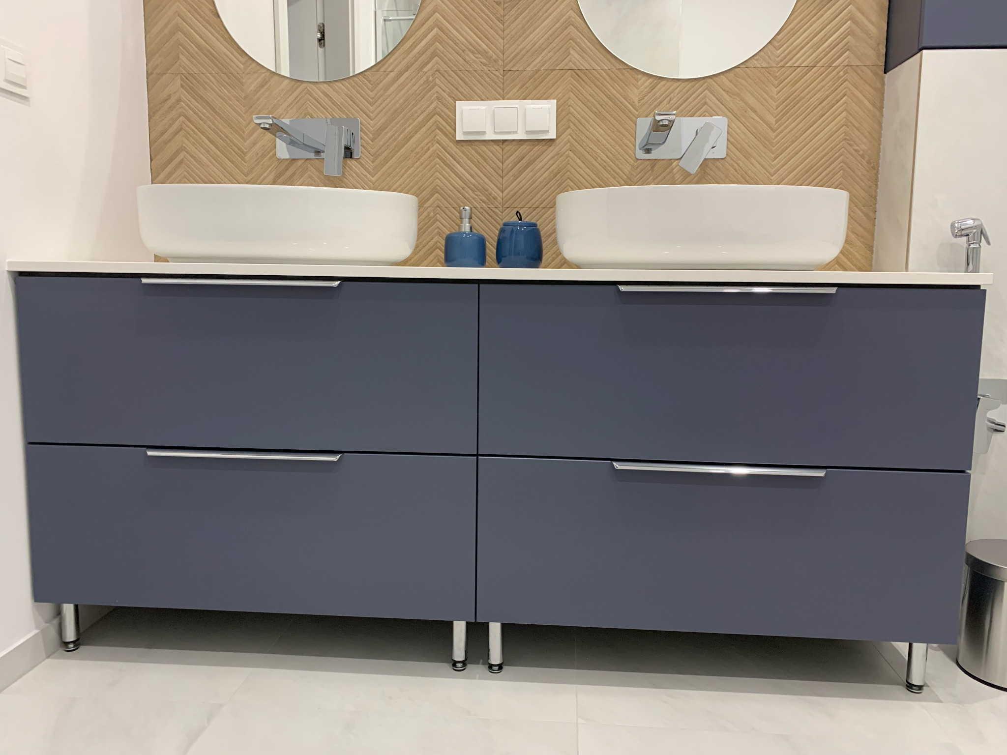 Niebieska szafka łazienkowa z dwiema umywalkami nablatowymi i chromowanymi bateriami, na tle ściany z wzorem jodełki. Nowoczesny design.