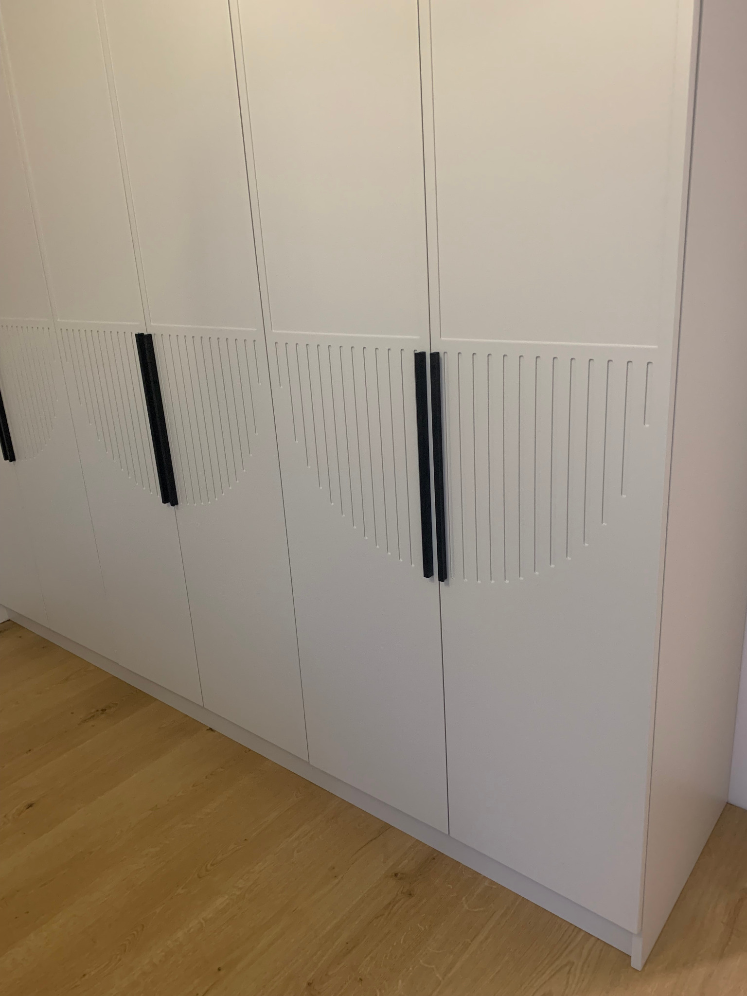 Biała szafa wnękowa z frezowanymi frontami i długimi, czarnymi uchwytami, widoczna na tle drewnianej podłogi. Minimalistyczny design.