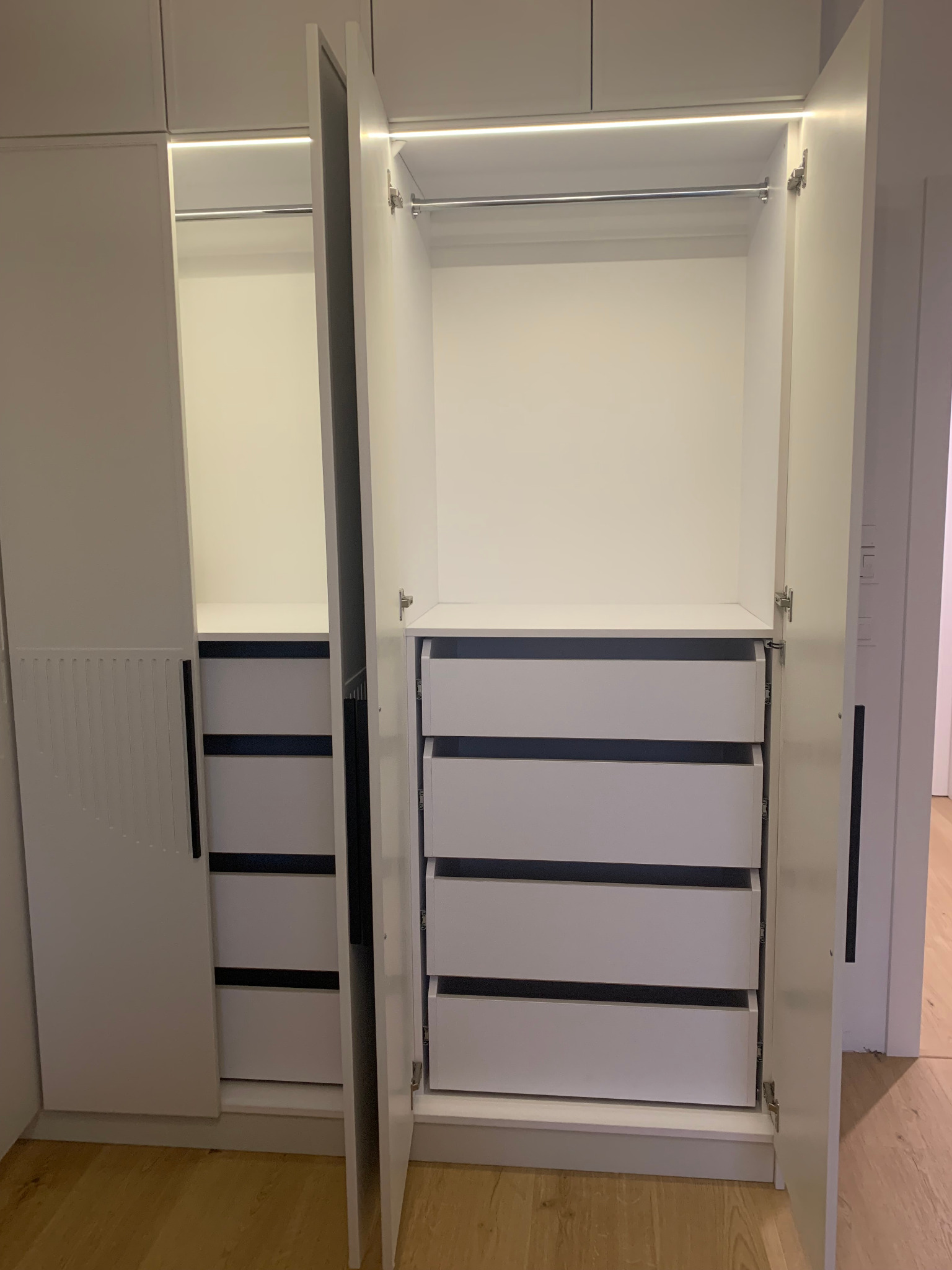 Biała szafa wnękowa z otwartymi drzwiami, ukazująca szuflady i półki. Oświetlenie LED wewnątrz. Minimalistyczny design, jasne wnętrze, drewniana podłoga.