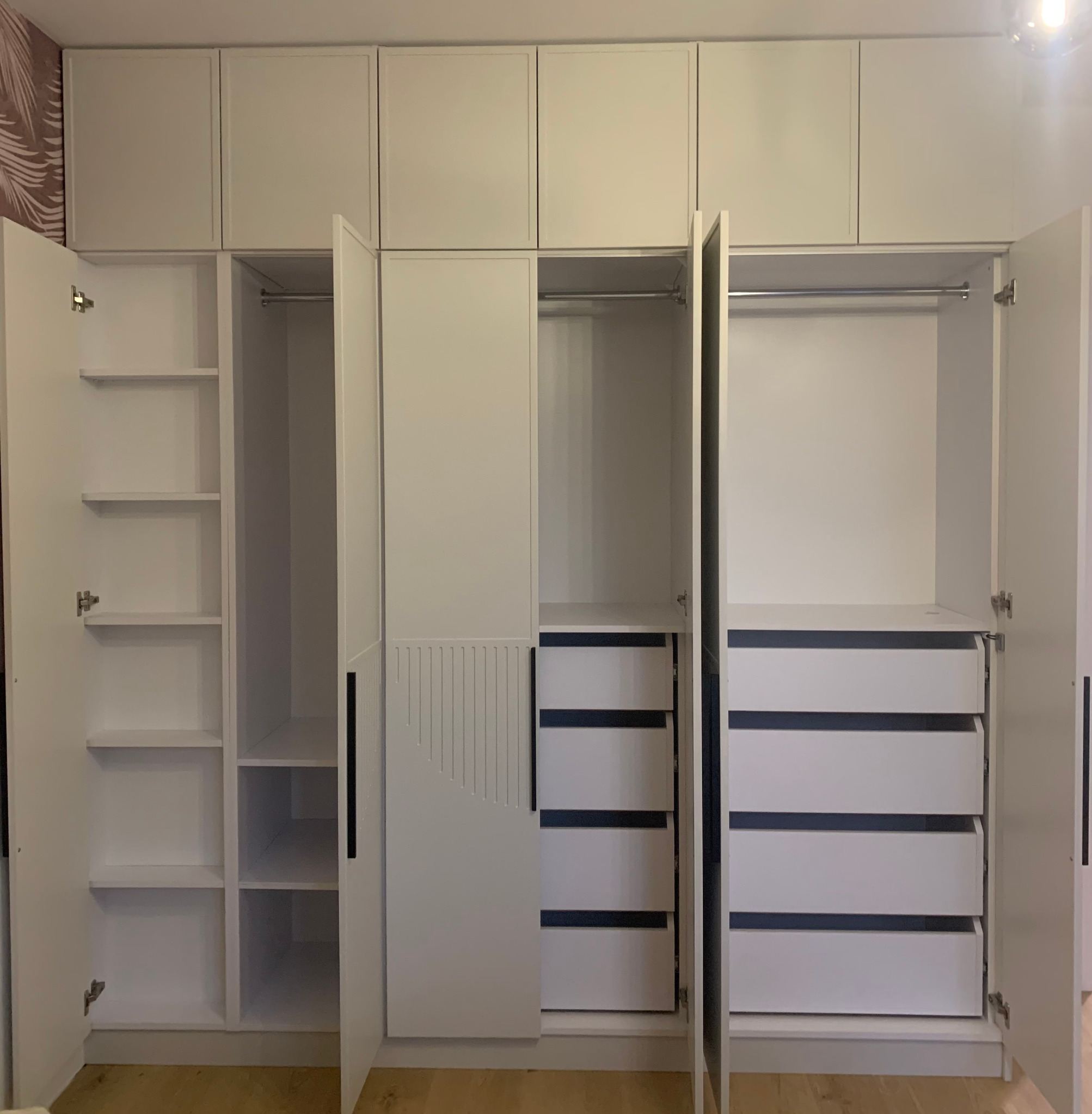 Biała szafa wnękowa z otwartymi drzwiami, ukazująca półki, drążek na ubrania i szuflady. Minimalistyczny design, czarne uchwyty, jasna podłoga.