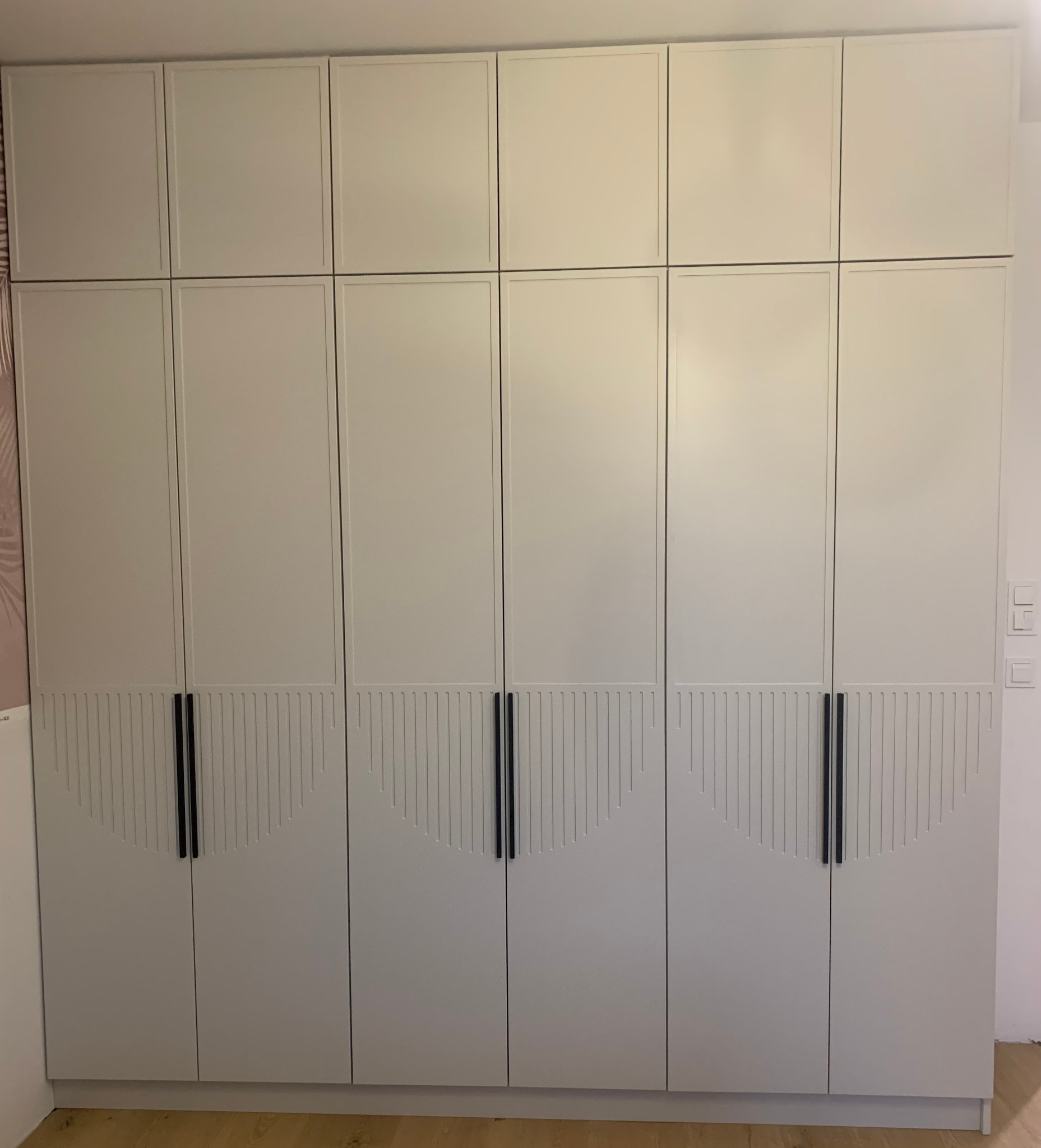 Biała szafa wnękowa z górnymi szafkami i dekoracyjnym frezowaniem na dolnych frontach, czarne, pionowe uchwyty, widok na całą ścianę.