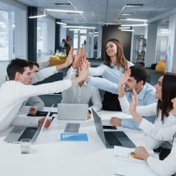 EfficienSync Business Solutions Tímea Hatos-Zatorska - Zespół biurowy świętuje sukces 'piątką' nad laptopami na białym stole w jasnym, nowoczesnym biurze z dużymi oknami i widocznym śniegiem na zewnątrz.