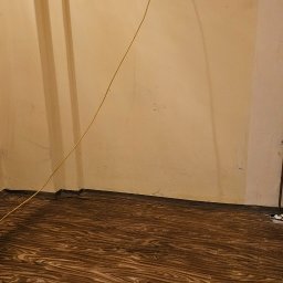 MAXI-ZIM DAMIAN KUCHARSKI - Ukończona podłoga z paneli imitujących drewno, widoczna ściana z gniazdkiem i wystającym kablem. Ujęcie z niskiego kąta, podkreślające wzór podłogi.