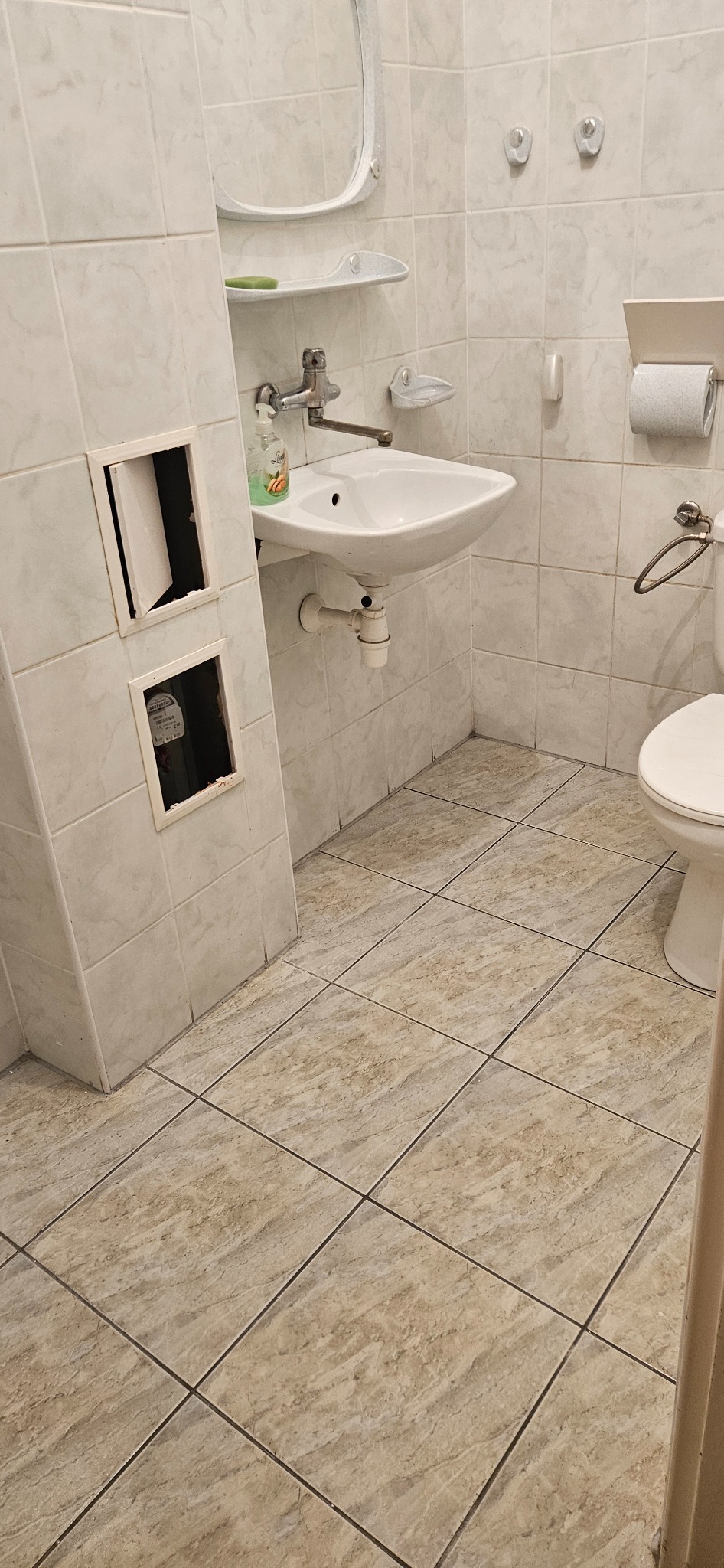 Łazienka z białą umywalką, toaletą i otwartymi otworami rewizyjnymi w obudowie. Widoczne płytki na ścianach i podłodze. Ujęcie z dołu, perspektywa lekko zniekształcona.