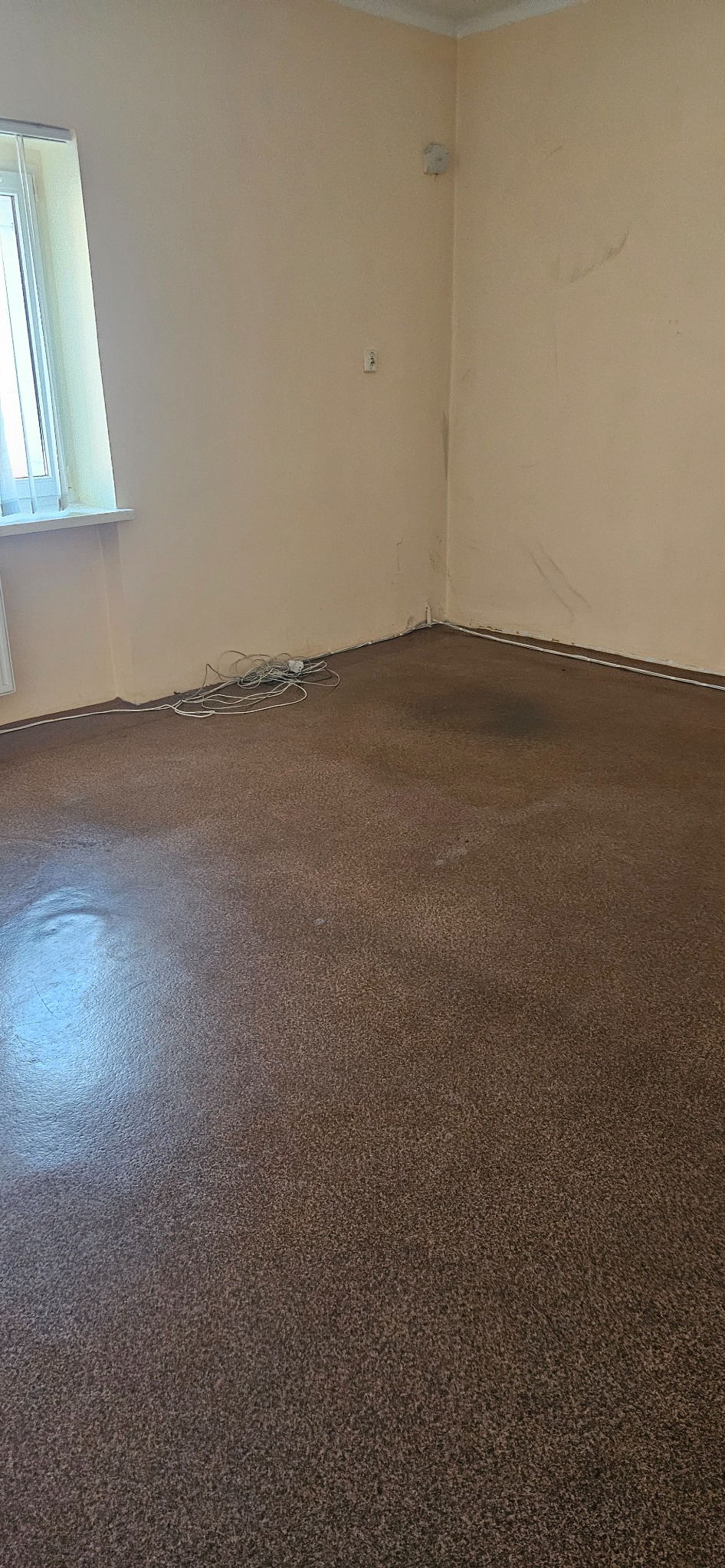 Pusty pokój z brązową podłogą, beżowymi ścianami i oknem po lewej. Widoczne ślady zabrudzeń na ścianach i podłodze. Na podłodze leżą zwinięte kable.