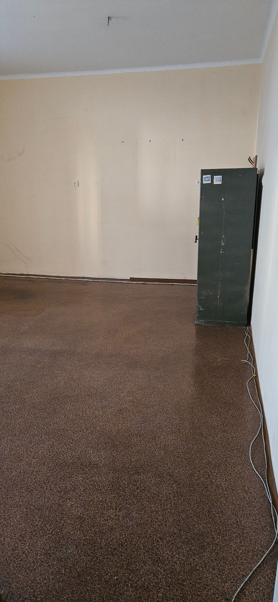 Pusta przestrzeń z brązową podłogą, beżowymi ścianami i ciemnozieloną szafą. Widoczne ślady po demontażu, sugerujące opróżnianie pomieszczenia.