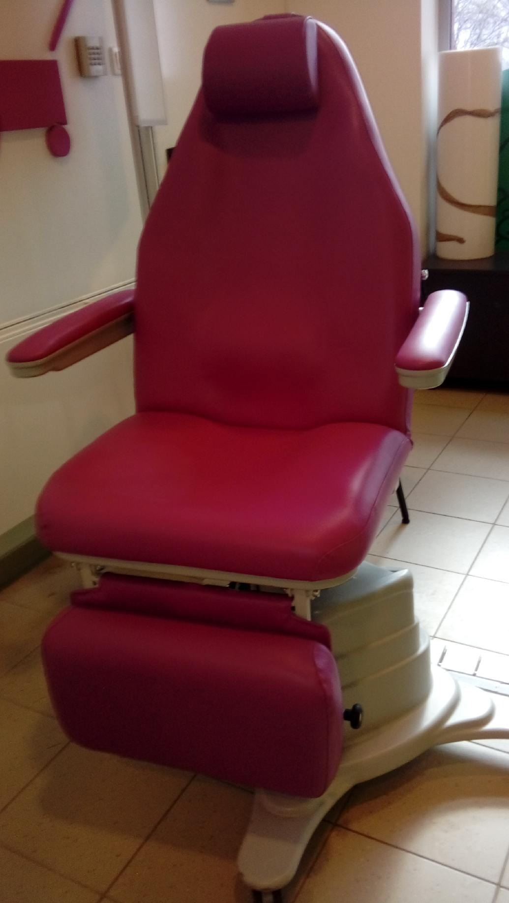 Różowe, tapicerowane krzesło medyczne z regulowanym podnóżkiem i zagłówkiem, ustawione na kafelkach. Widoczne elementy metalowe i plastikowe.