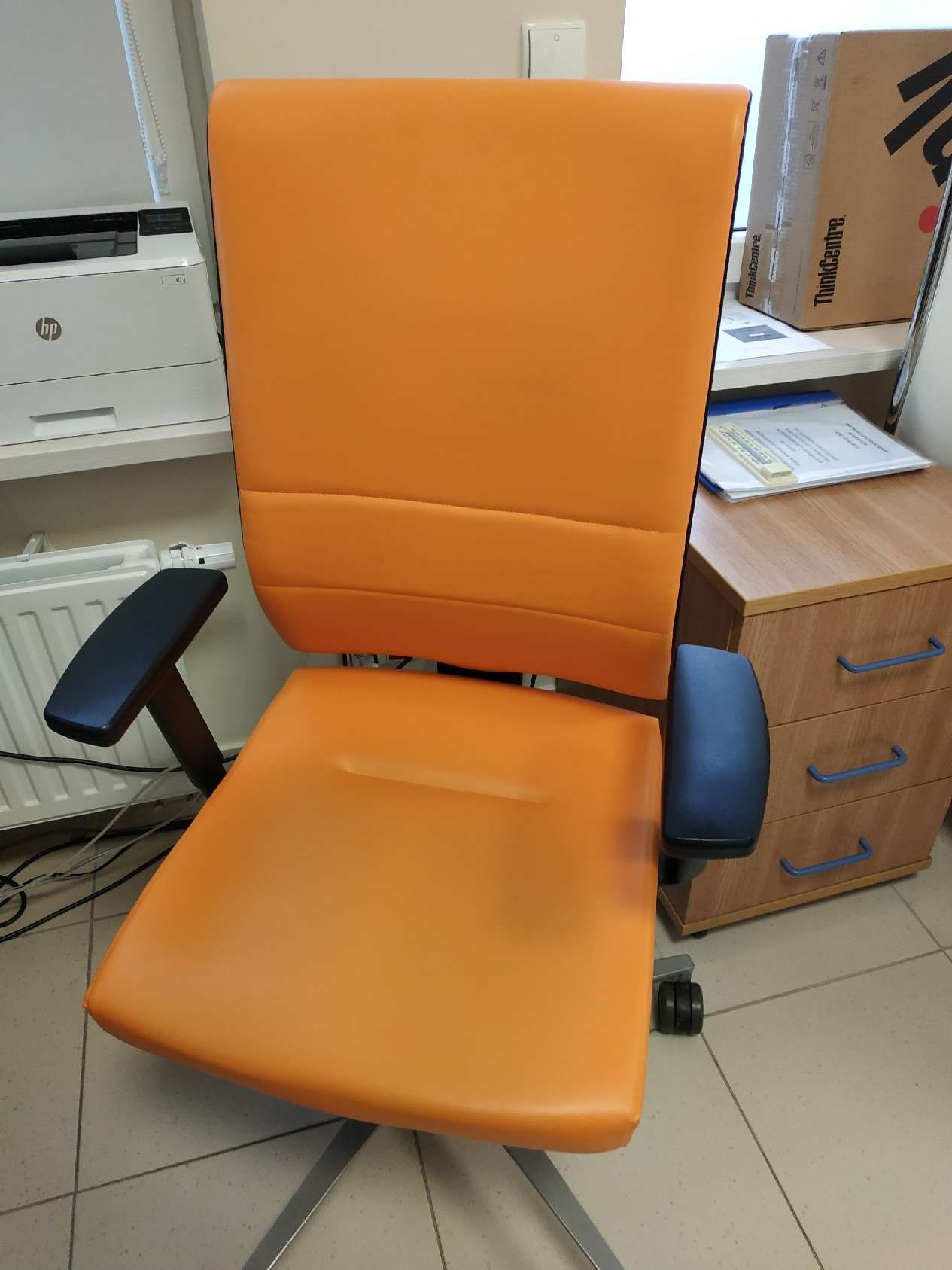 Pomarańczowe krzesło biurowe z czarnymi podłokietnikami na tle biura. Widoczny fragment biurka i drukarki. Krzesło na kółkach, z wysokim oparciem.