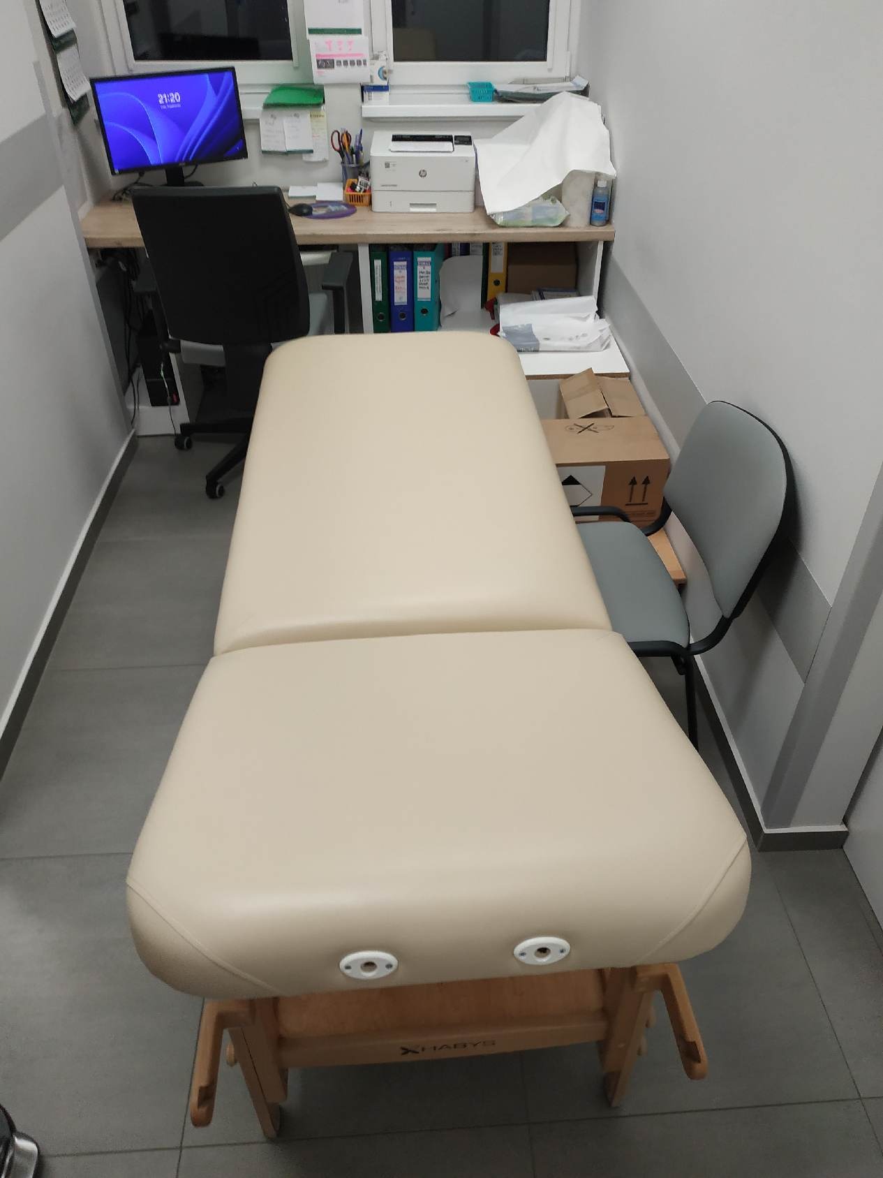 Beżowy, tapicerowany stół do masażu z drewnianą podstawą w jasnym gabinecie. Widoczny monitor, biurko i krzesło w tle. Minimalistyczny wystrój wnętrza.