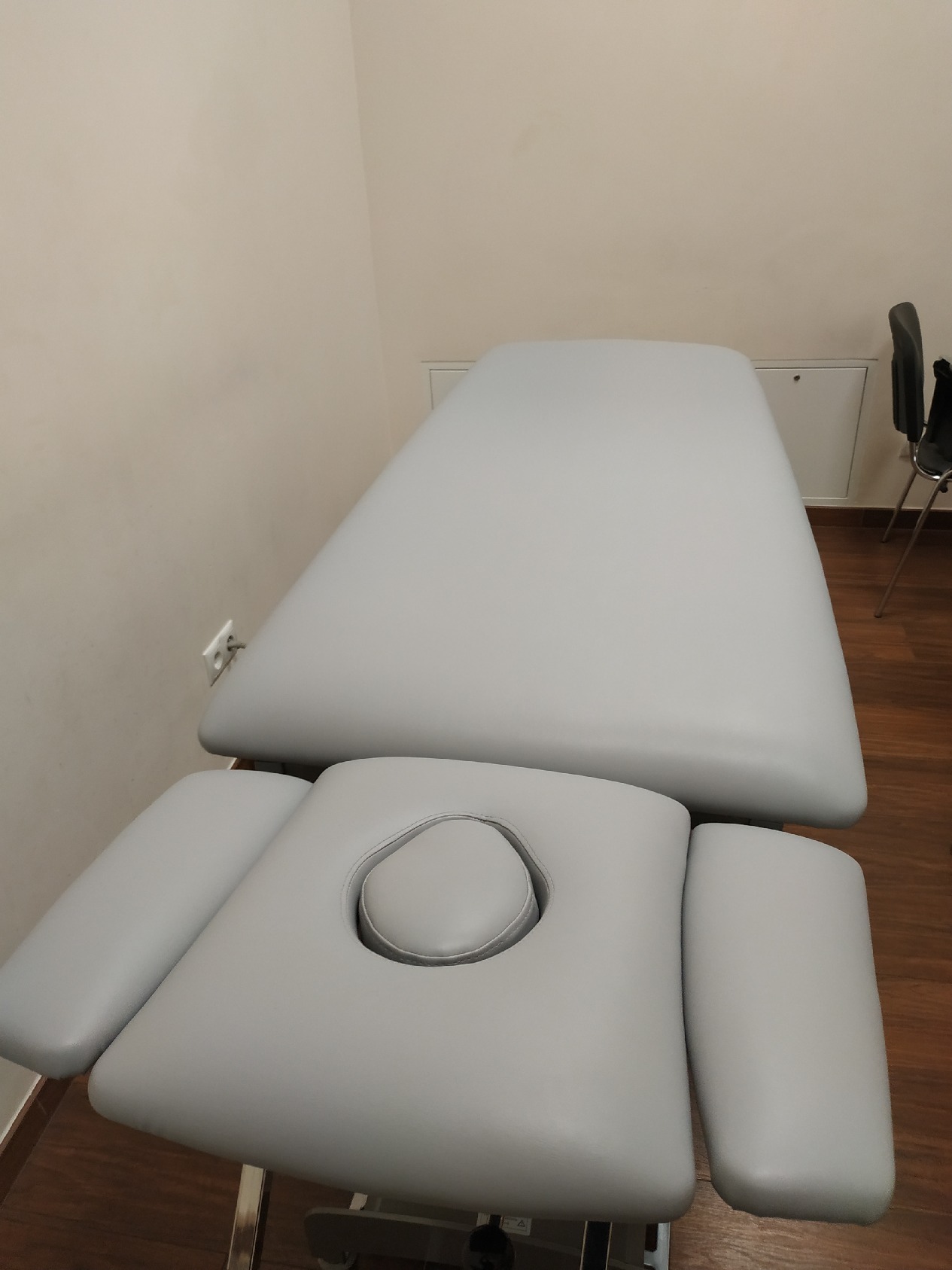 Szary, tapicerowany stół do masażu z otworem na twarz i regulowanymi podłokietnikami, ustawiony w jasnym pomieszczeniu z drewnianą podłogą.
