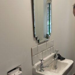 zibexpro - Biała umywalka z baterią retro i lustrem art deco na ścianie z nowo ułożonymi białymi płytkami. Widoczne krzyżyki dystansowe.