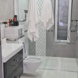 zibexpro - Nowoczesna łazienka z szarymi płytkami, wiszącą toaletą i umywalką z szafką. Na ścianie dekoracyjne płytki z wzorem rombów. Widoczne ręczniki i okno z żaluzjami.