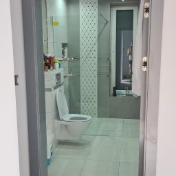 zibexpro - Nowoczesna łazienka z toaletą typu Geberit, widoczna przez szarą futrynę drzwiową. Białe płytki podłogowe i dekoracyjne panele ścienne. W tle okno z odbiciem osoby.