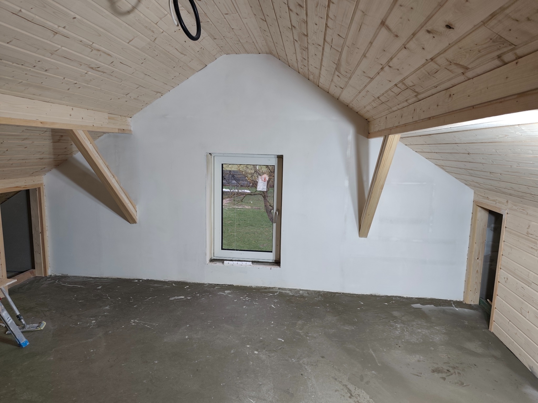Poddasze ze skośnym drewnianym sufitem i oknem. Ściany pomalowane na biało, surowa podłoga. Widoczne drewniane elementy konstrukcyjne i drabina malarska.