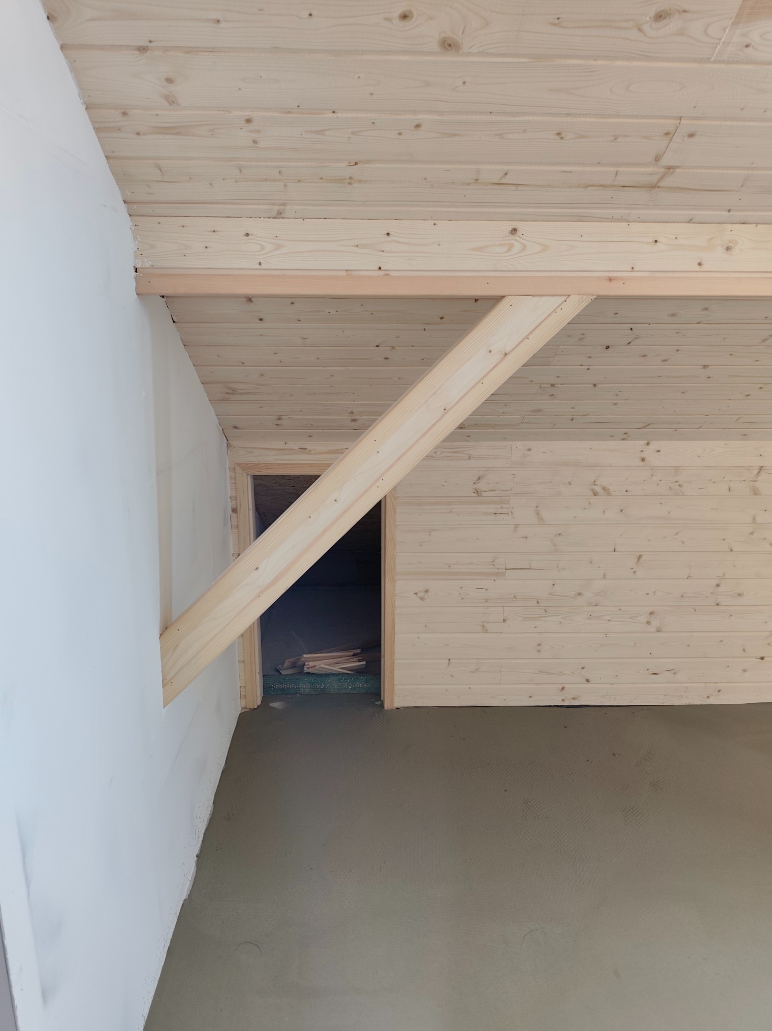 Poddasze z drewnianą boazerią na ścianach i suficie, surowy beton na podłodze, widoczna konstrukcja nośna i wejście do innego pomieszczenia.