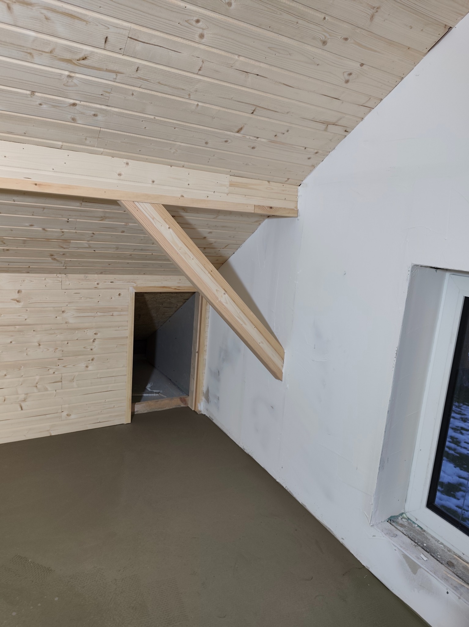 Poddasze po wylewce, ściany częściowo obłożone boazerią, widoczne drewniane elementy konstrukcyjne i okno. Szare wykończenie podłogi, surowe ściany czekają na dalsze prace.