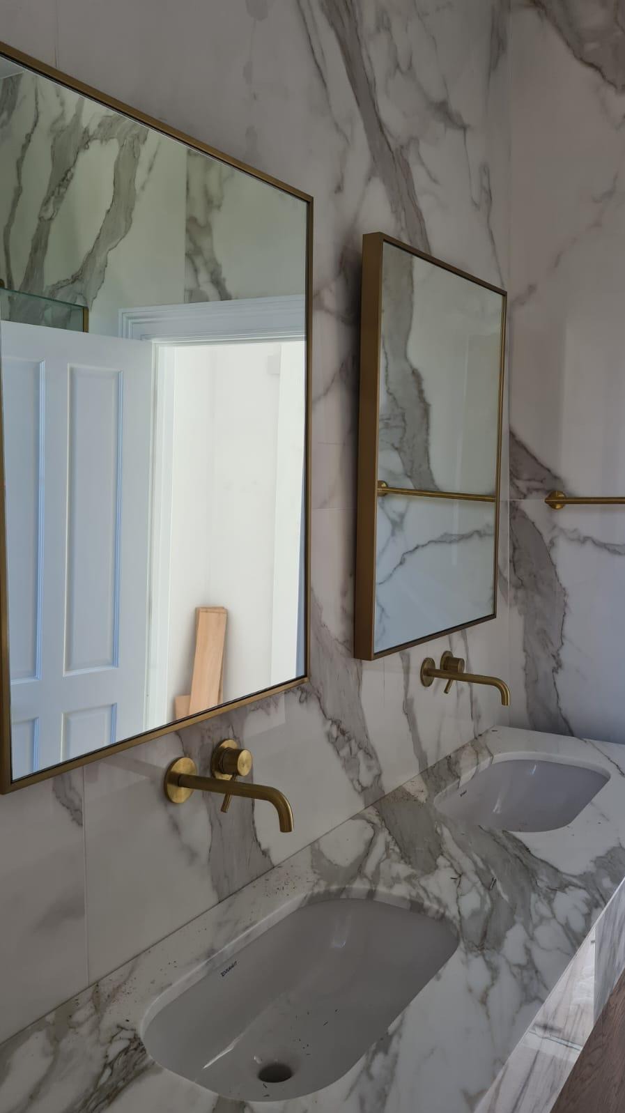 Elegancka łazienka z marmurowymi płytkami, złotymi bateriami i lustrami w złotych ramach. Widoczne dwie umywalki i fragment otwartych drzwi w odbiciu lustra.