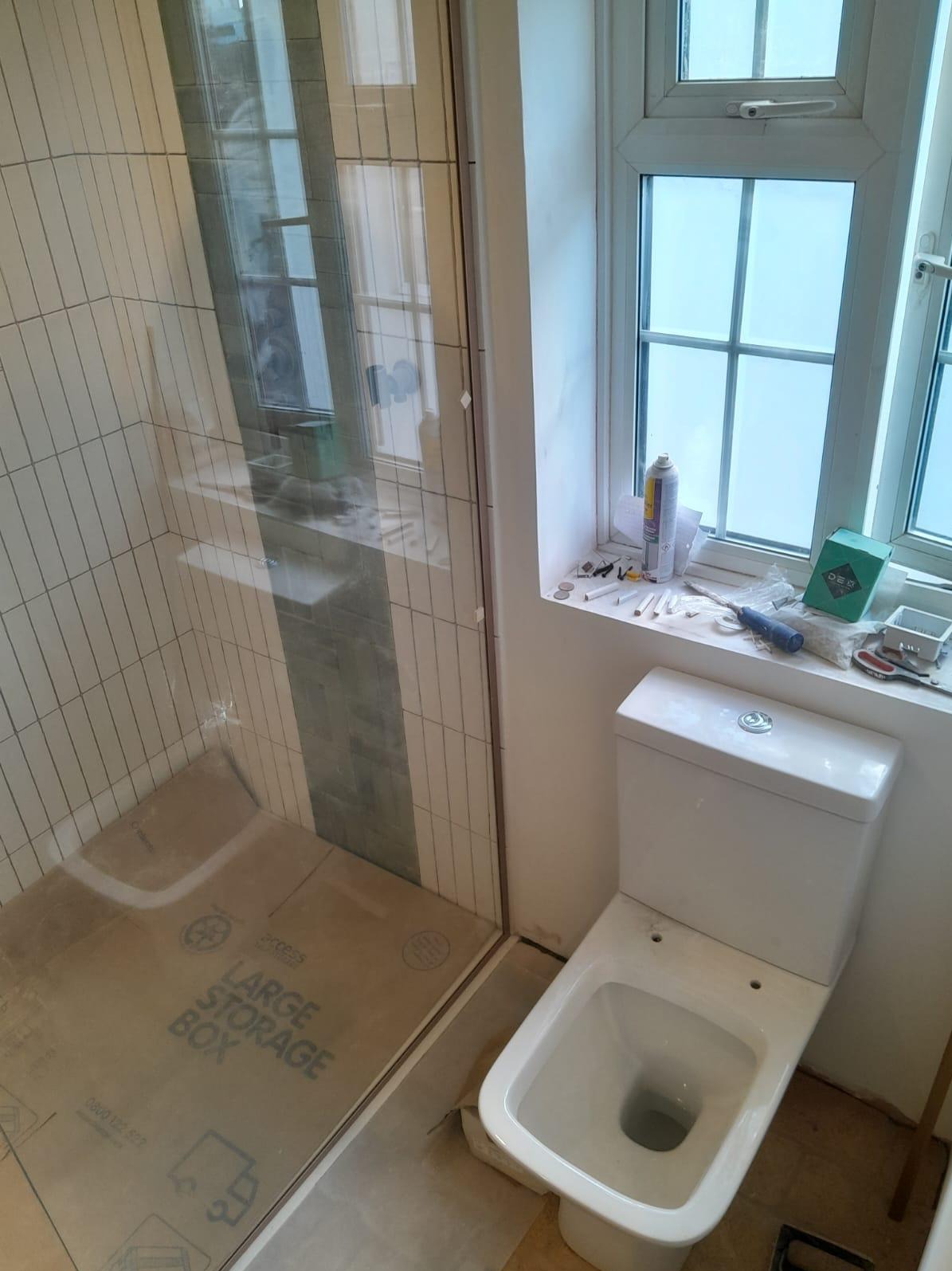 Nowa kabina prysznicowa z białymi płytkami i szklanymi drzwiami, obok toaleta i okno z akcesoriami na parapecie. Widok na łazienkę w trakcie wykańczania.