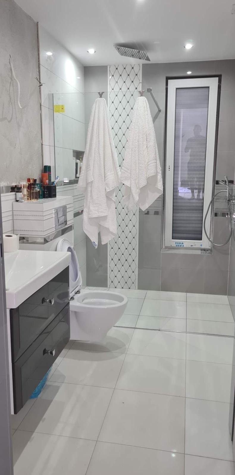 Nowoczesna łazienka z szarymi płytkami, wiszącą toaletą i umywalką z szafką. Na ścianie dekoracyjne płytki z wzorem rombów. Widoczne ręczniki i okno z żaluzjami.