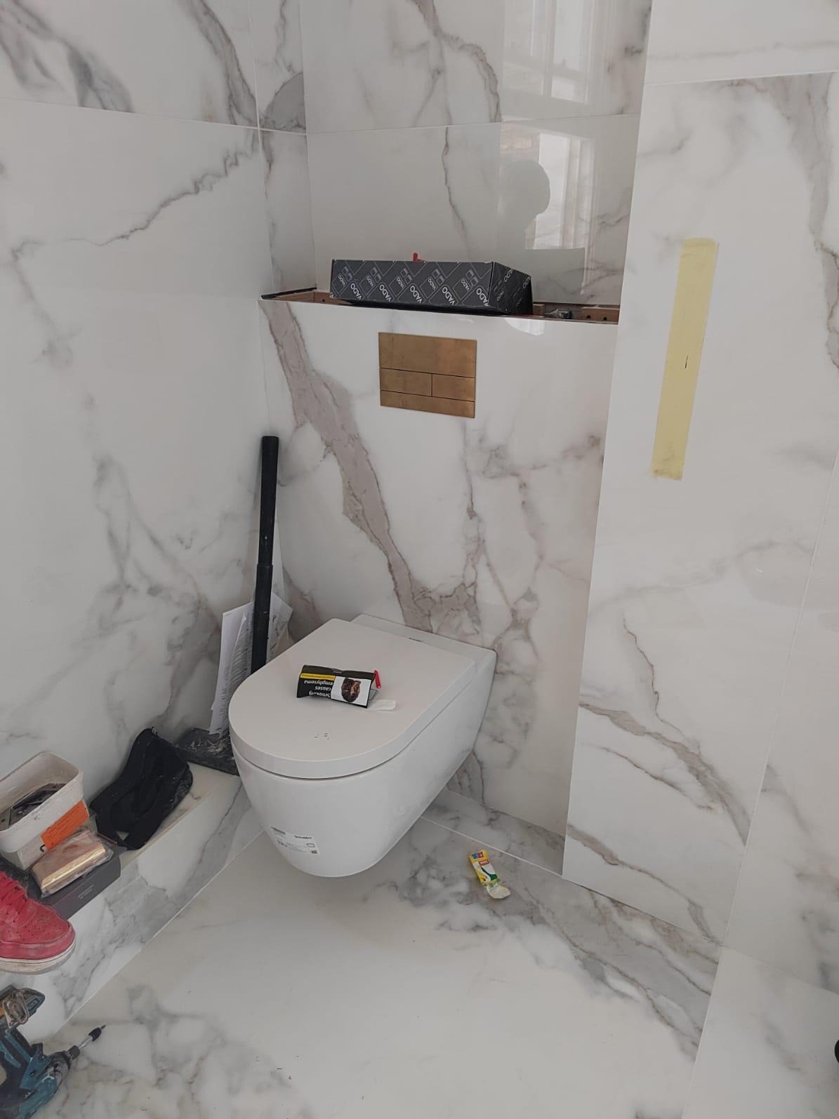 Biała toaleta wisząca w łazience wykończonej płytkami imitującymi marmur. Widoczna złota armatura i akcesoria. Na sedesie leży paczka papierosów.