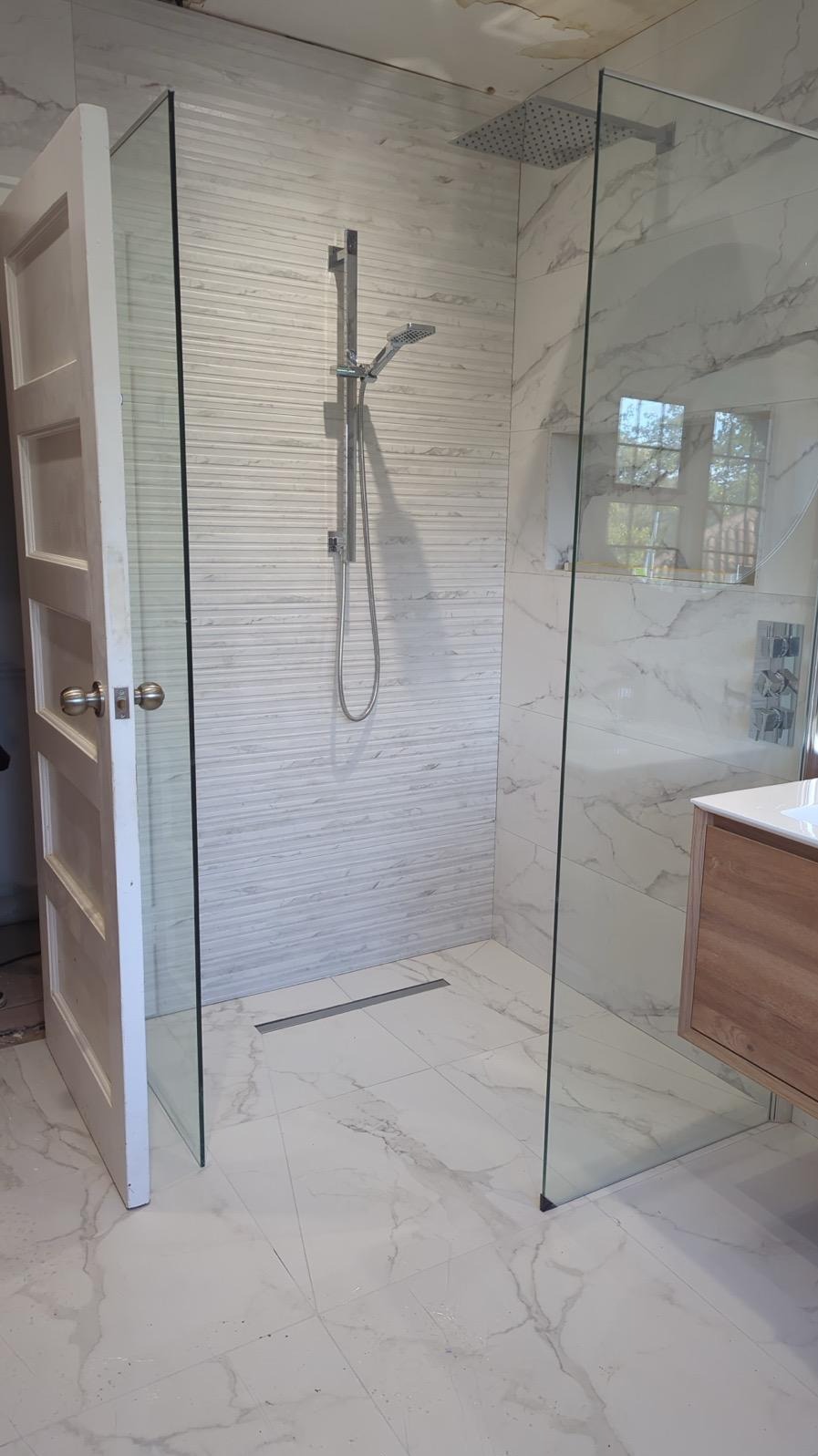 Nowoczesna kabina prysznicowa walk-in z odpływem liniowym, wykończona płytkami imitującymi marmur i drewnianą szafką. Widoczna otwarta, biała, drewniana rama drzwi.